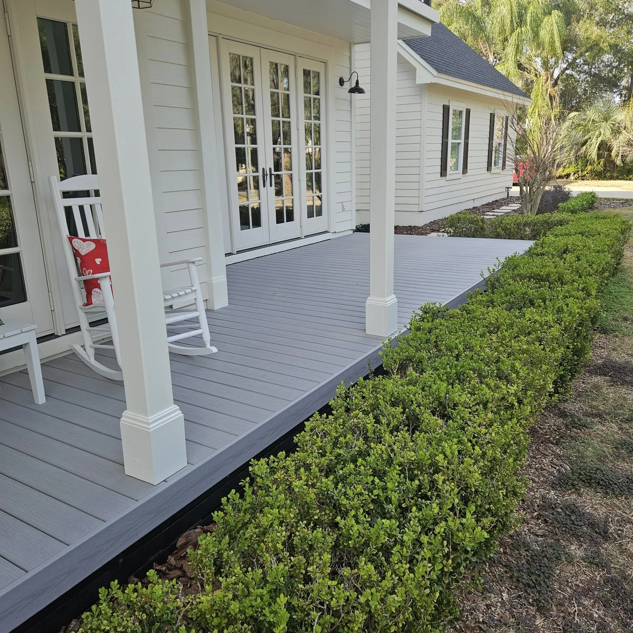 A backyard porch with a gray composite deck, white siding house, and glass double doors. There are green bushes, a small tree, shaded with palm trees in the background.