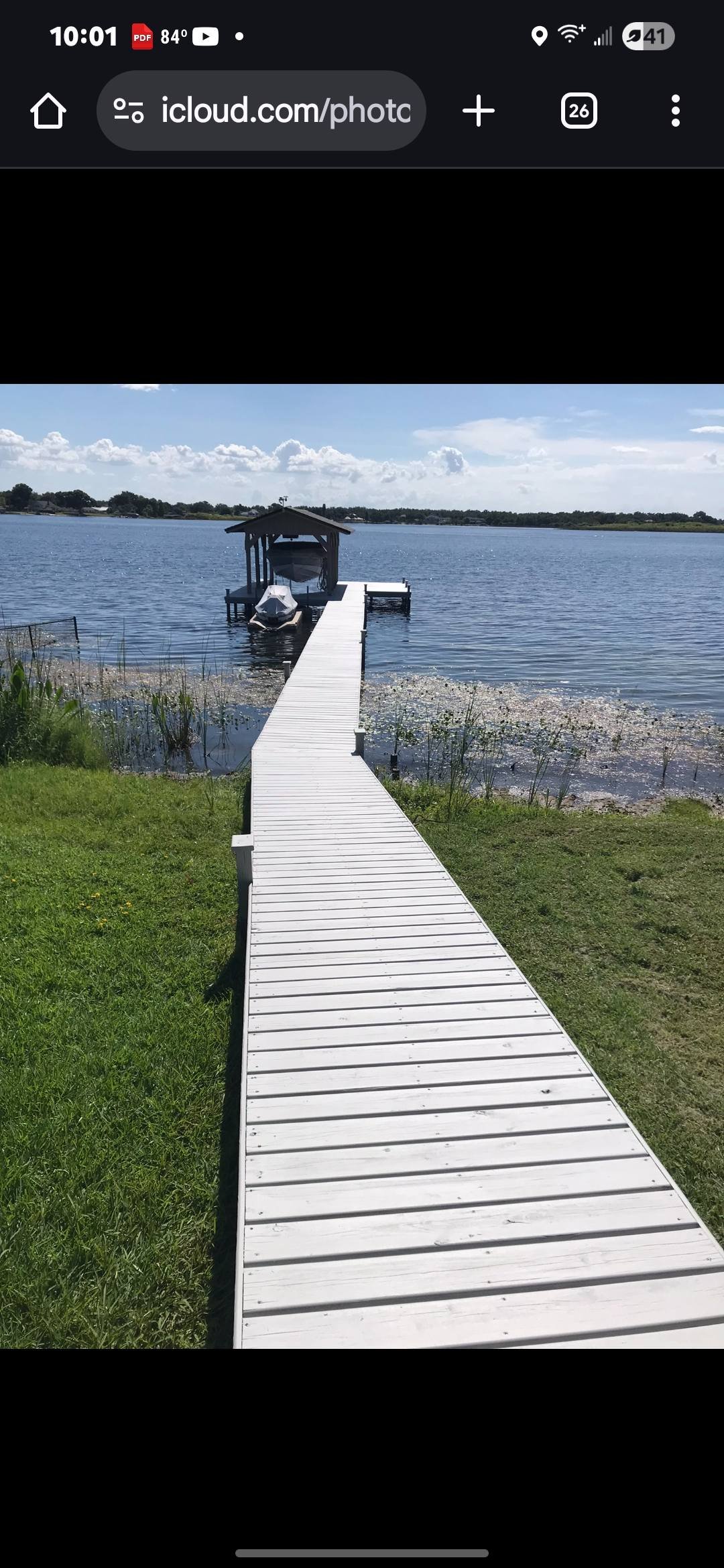A white wooden dock extending over a calm lake, with a boat covered by a canopy and another boat docked at the end of the pier. Green grass and plants line the shoreline, with a partly cloudy sky overhead.