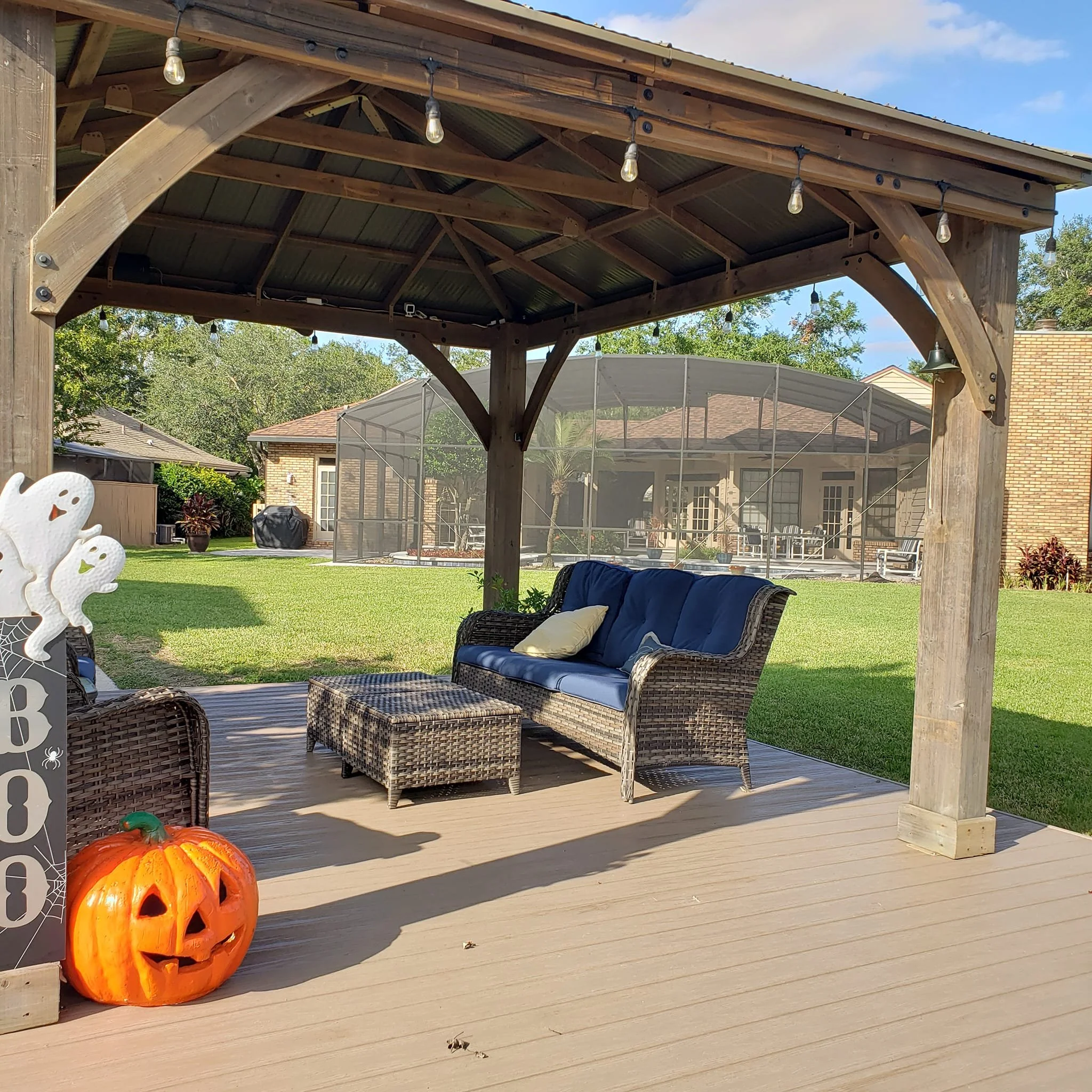 A decorated outdoor patio area with wicker furniture, string lights hanging from a wooden pergola, and a large orange pumpkin with a carved face on the ground in front of the patio. The background includes a backyard with green grass, trees, and a sc