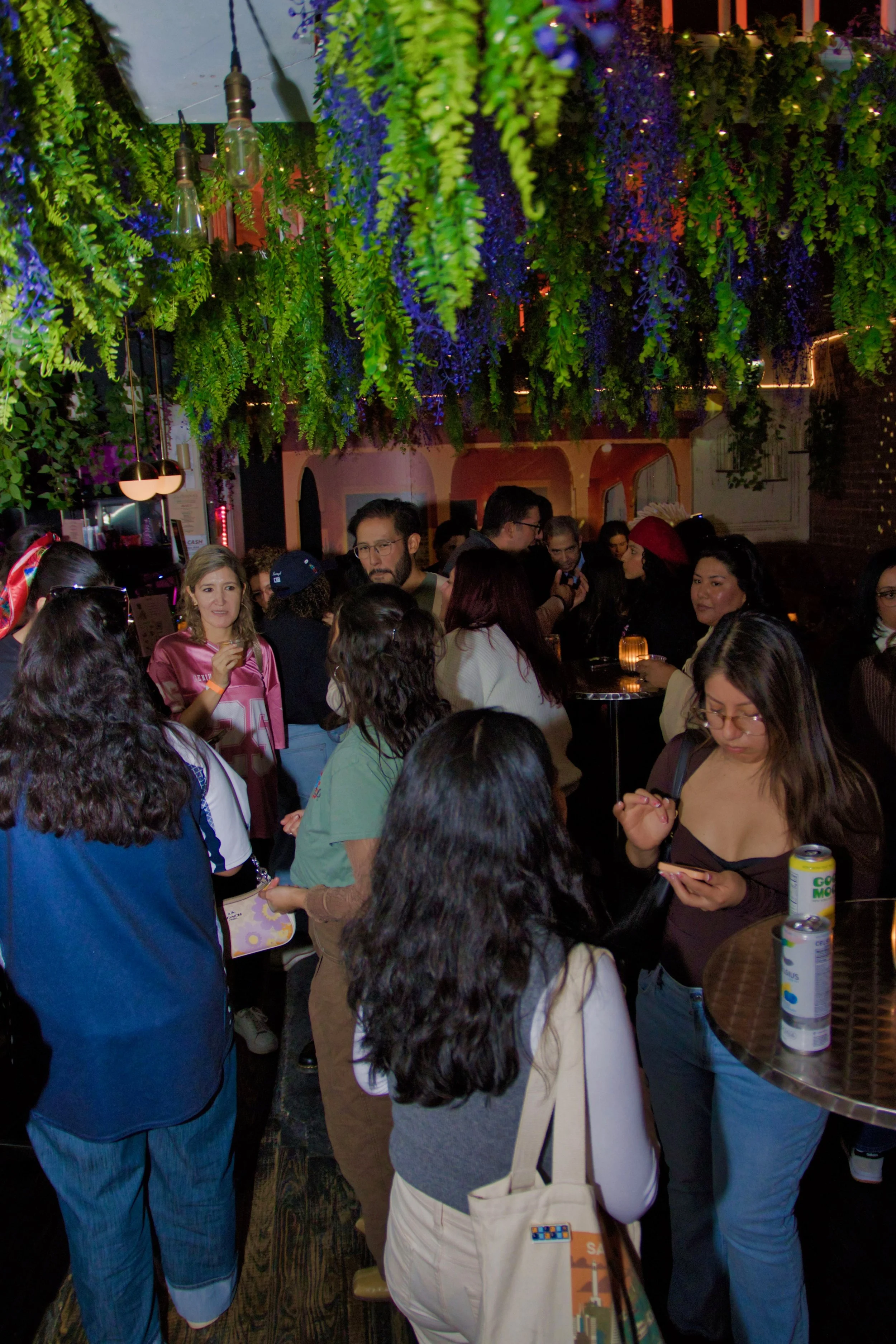 Crowd of people gathered in a dimly lit, festive indoor space decorated with hanging greenery and purple flowers, with string lights overhead.