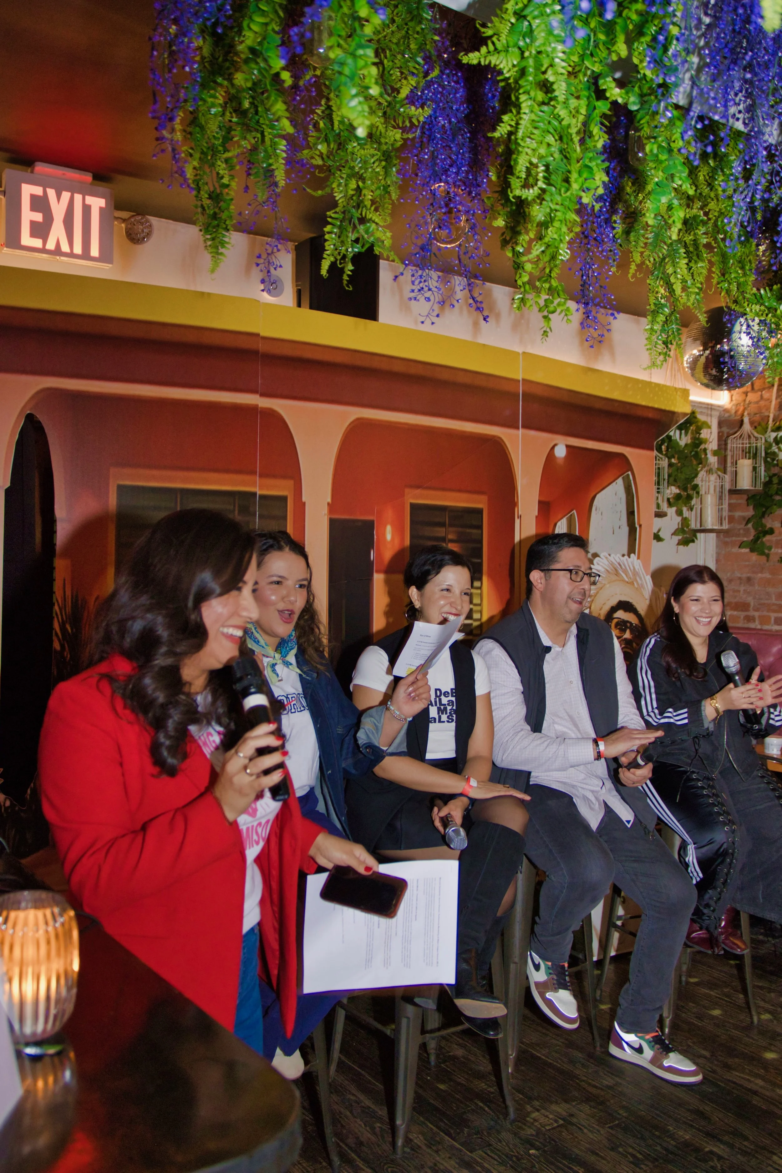 Group of five people sitting and laughing in a restaurant or bar, some holding microphones, with a decorated ceiling with hanging plants and a disco ball.