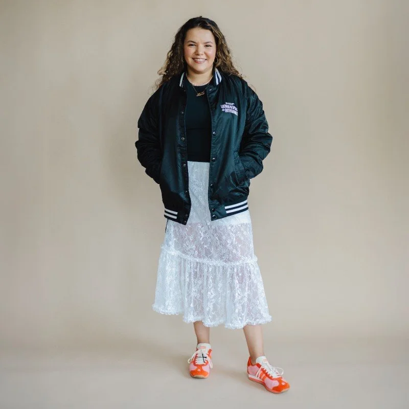 Young woman smiling, wearing a black jacket, long white lace skirt, and sneakers, standing against a neutral background.