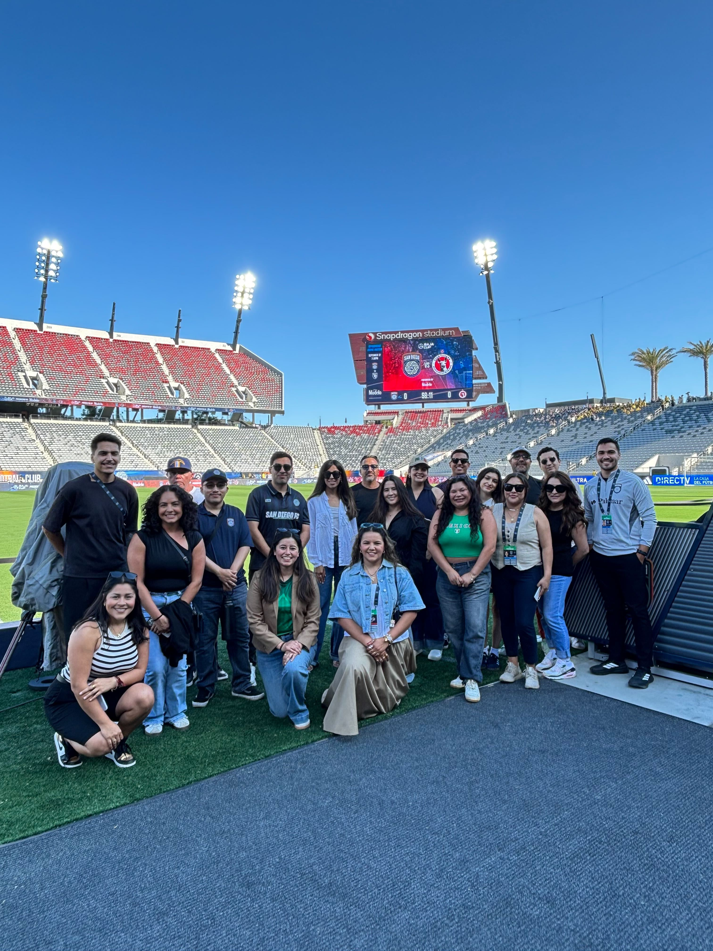 Group of people standing on a grass field inside Snapdragon Stadium with a large digital scoreboard and stadium seating in the background on a clear, sunny day.