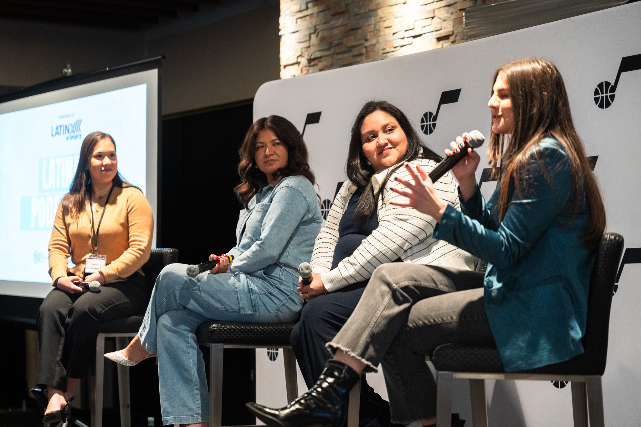 Four women seated on stage participating in a panel discussion at a Latino event, with a large screen and branding behind them.