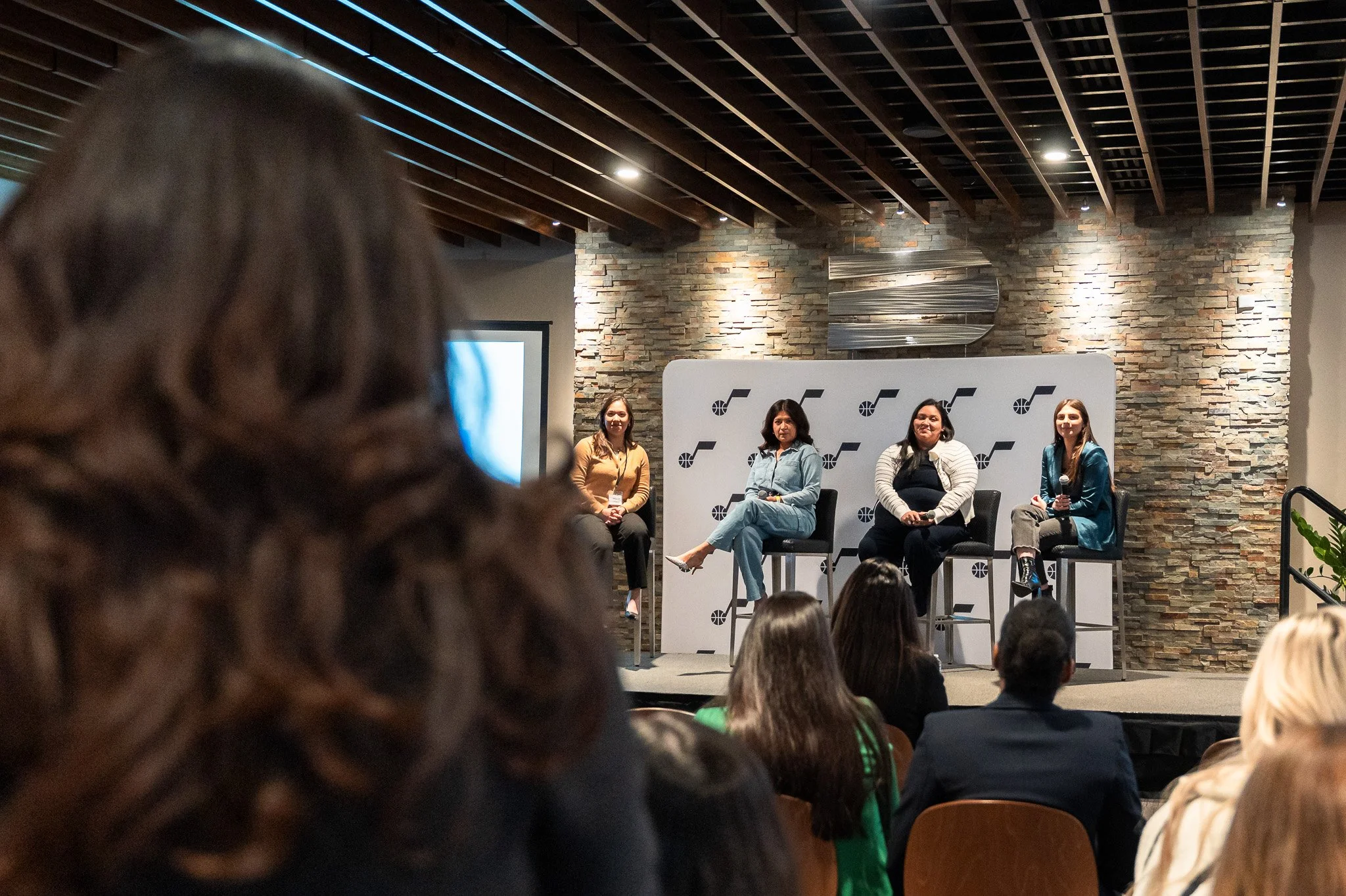 Four women seated on a stage during a panel discussion at a conference, with an audience in front of them and a woman with curly hair in the foreground.