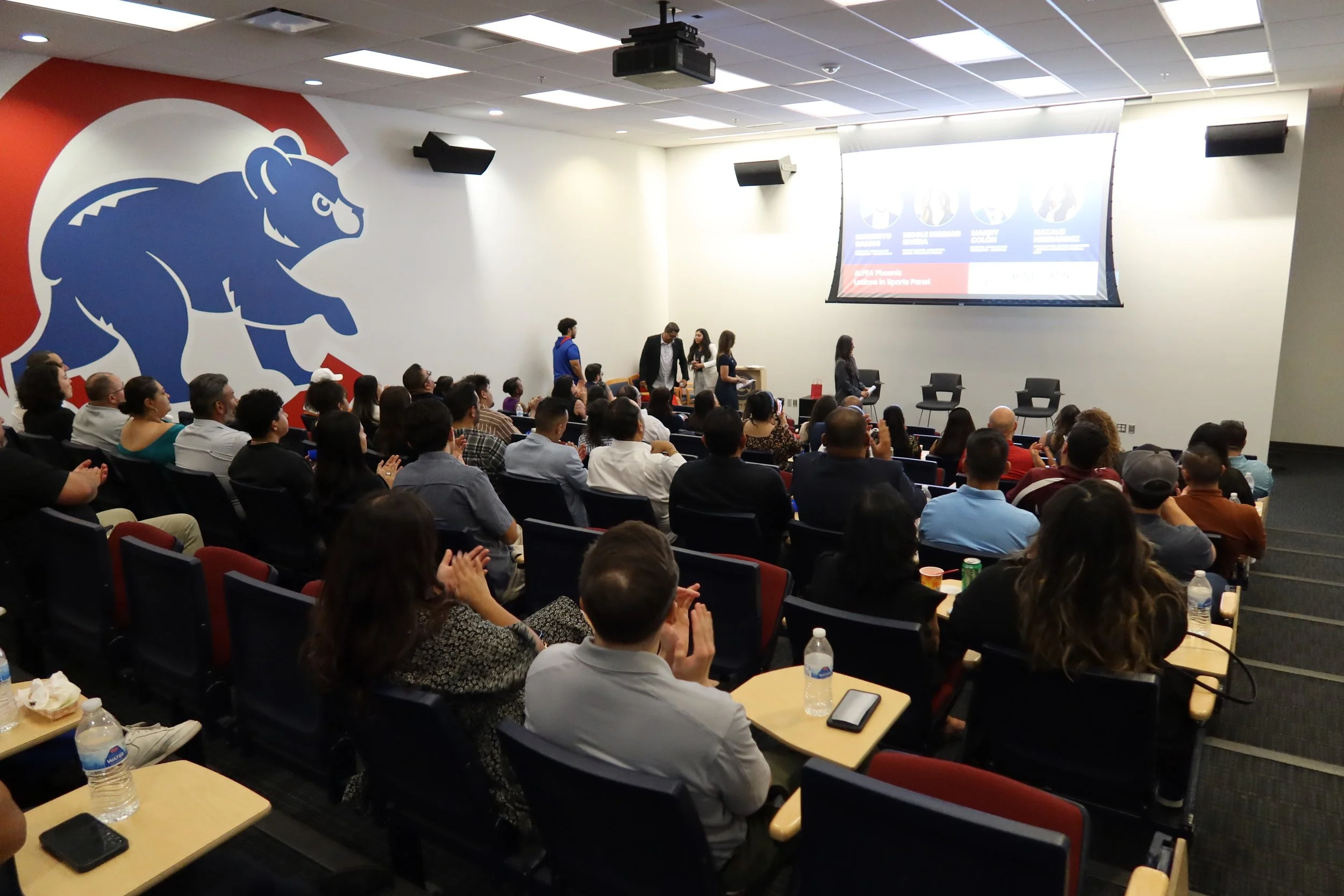 A large audience in an auditorium watching a presentation. The room has a large blue and white bear logo on the wall, and a projection screen displaying slides. Some people are clapping, and there are tables with water bottles and phones in front of some seats.