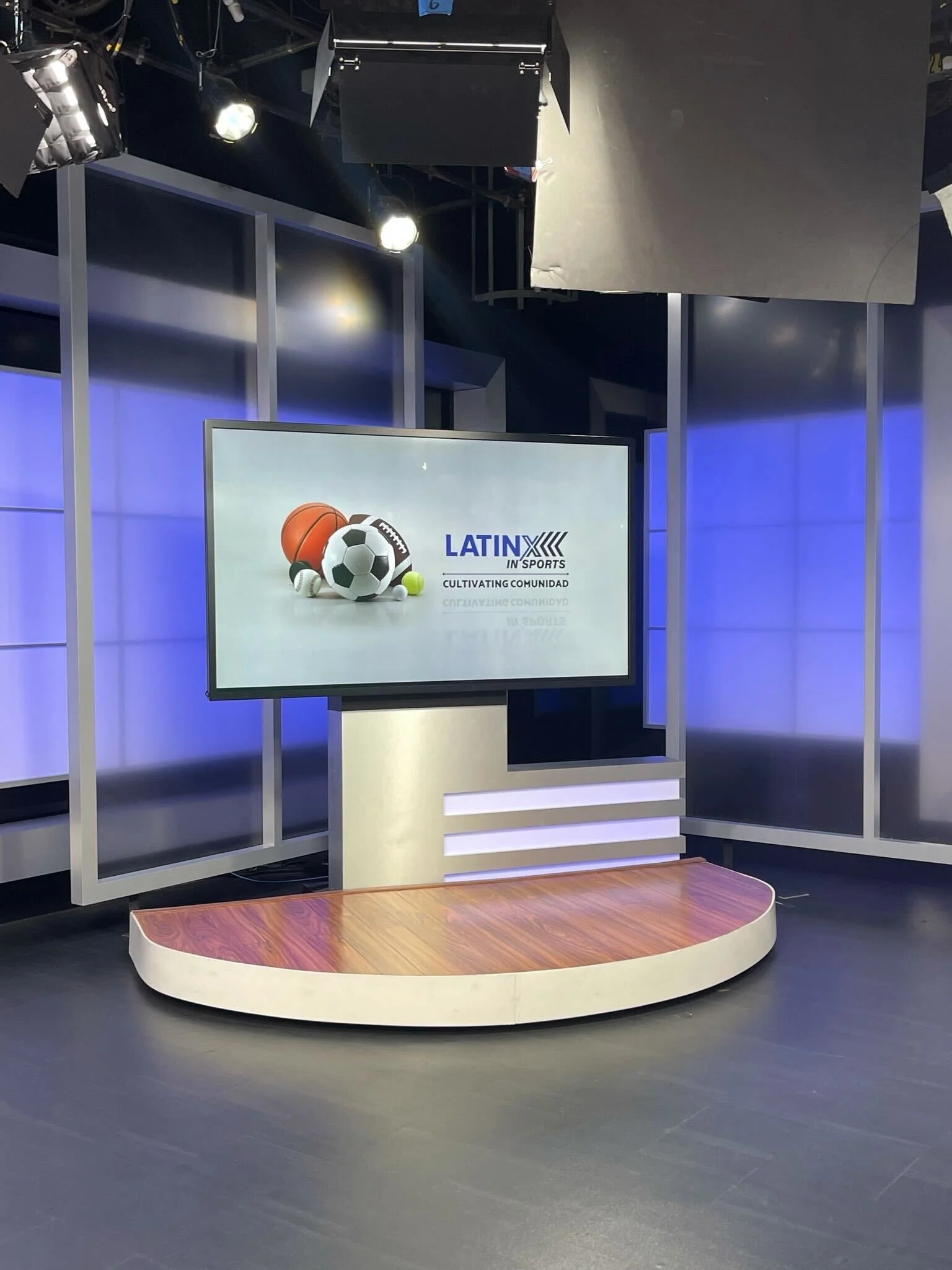 Television studio with a large screen displaying sports equipment and the text 'Latinx in Sports' and 'Cultivating Comunidad', a wooden platform, studio lights, and a blue-lit background.