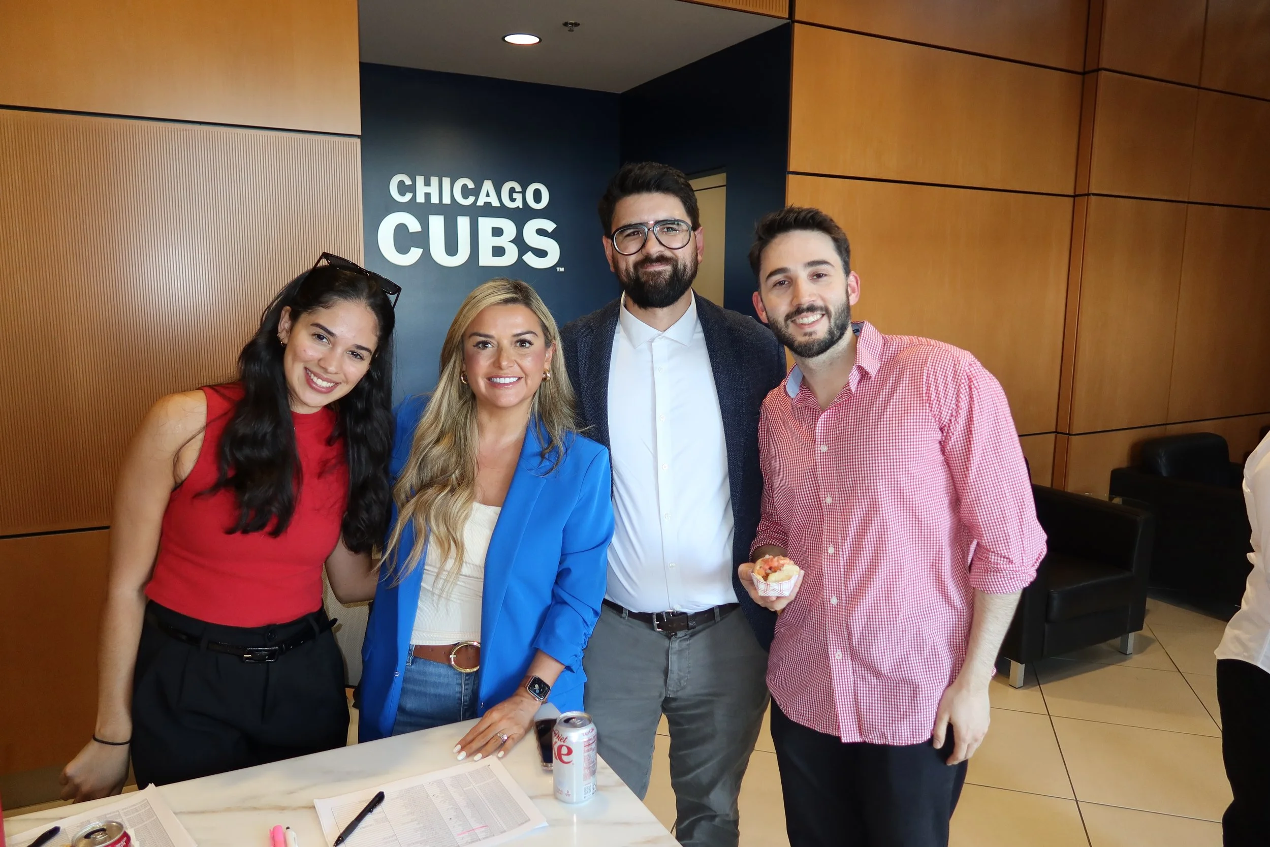 Four people standing together in front of a wall with a 'Chicago Cubs' sign, smiling and posing for the photo, with two women and two men, one woman has long dark hair, another blonde, and the men have beards. One man is holding a cupcake, and there are drinks and papers on the table in front of them.