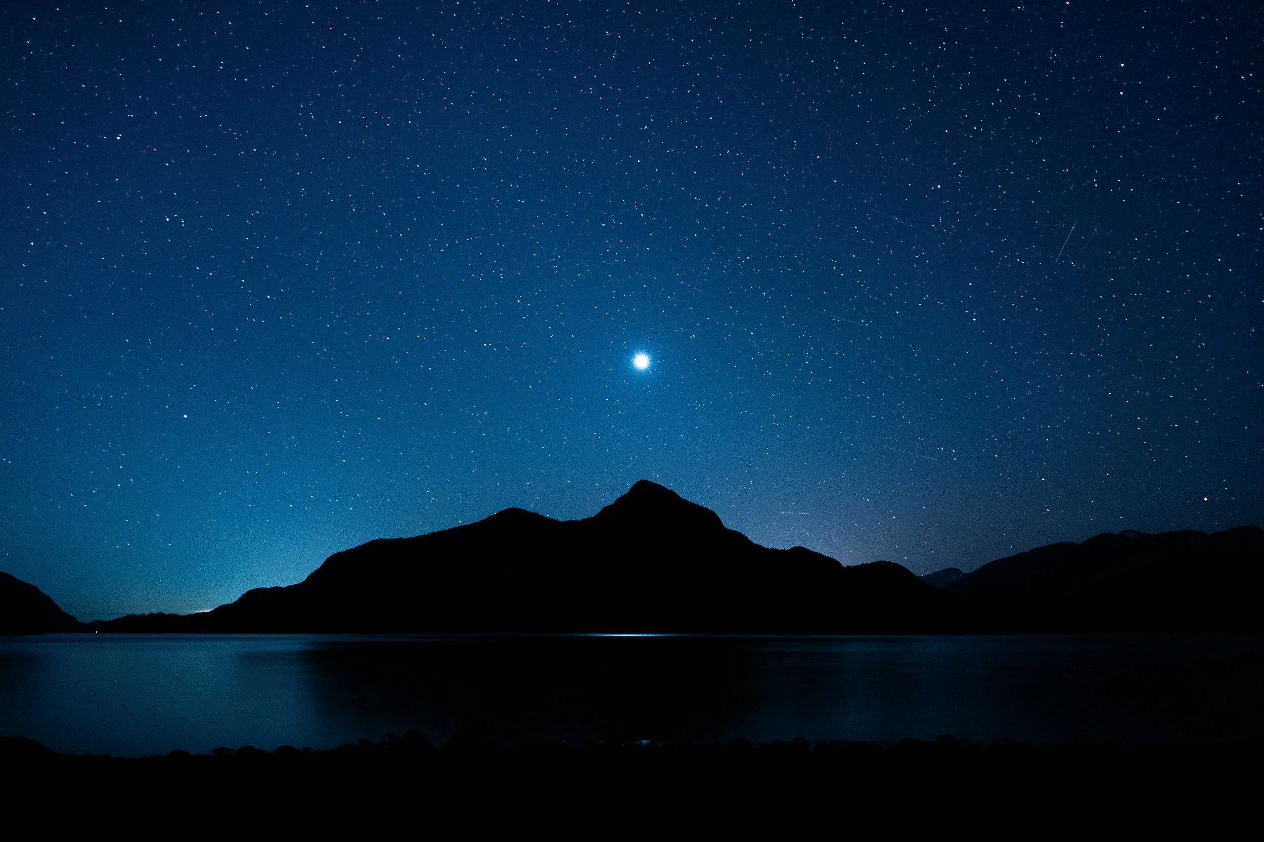 Nighttime landscape with silhouettes of mountains and a lake, star-filled sky with a bright planet or star in the center.