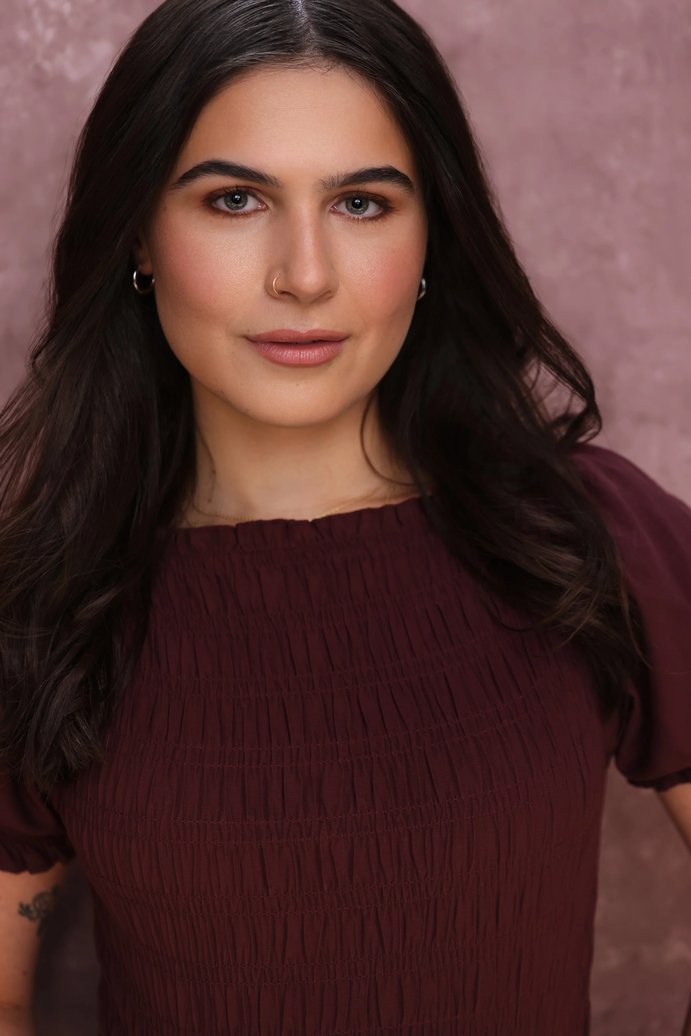 Close-up portrait of a young woman with dark hair and blue eyes, wearing a maroon blouse and hoop earrings, against a pinkish background.