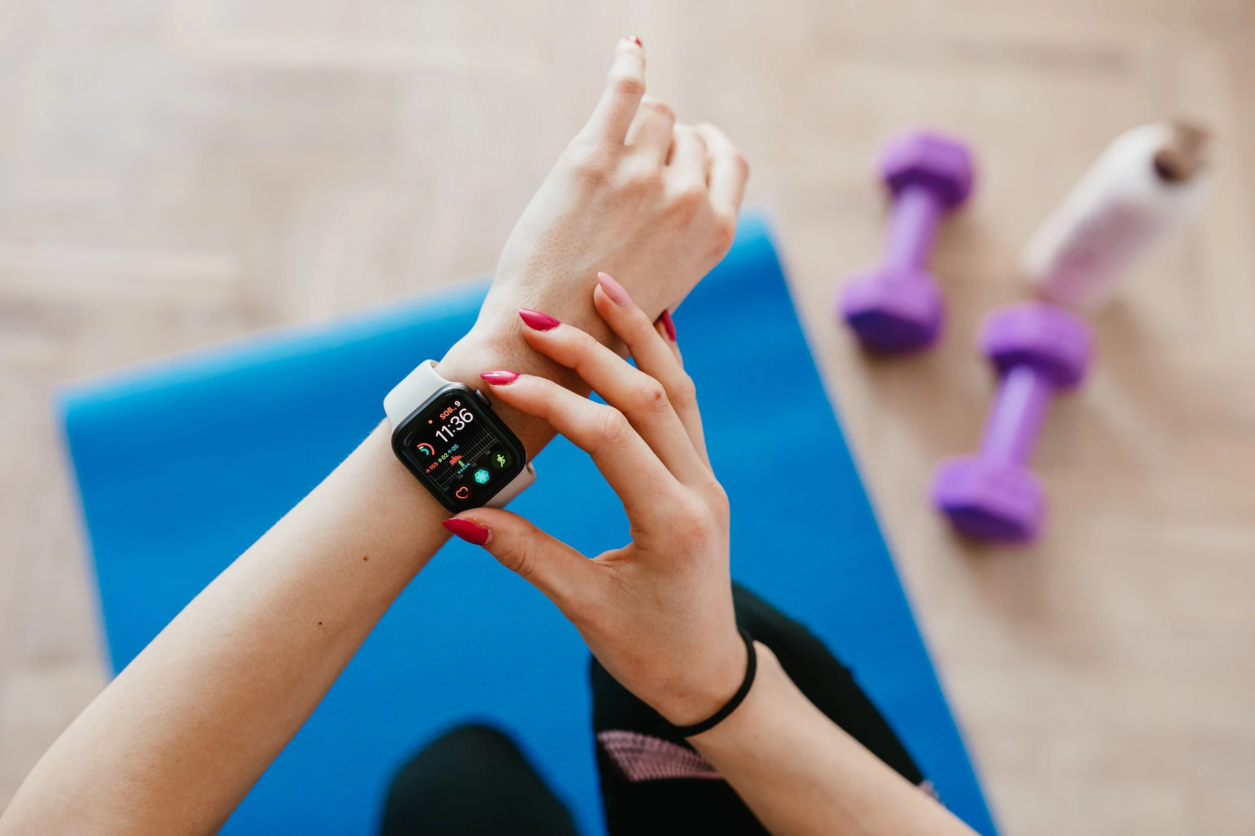 Woman checking an Apple Watch fitness tracker on a blue mat with purple dumbbells nearby representing workout progress tracking and daily fitness goals. Photo by Karola G on Pexels