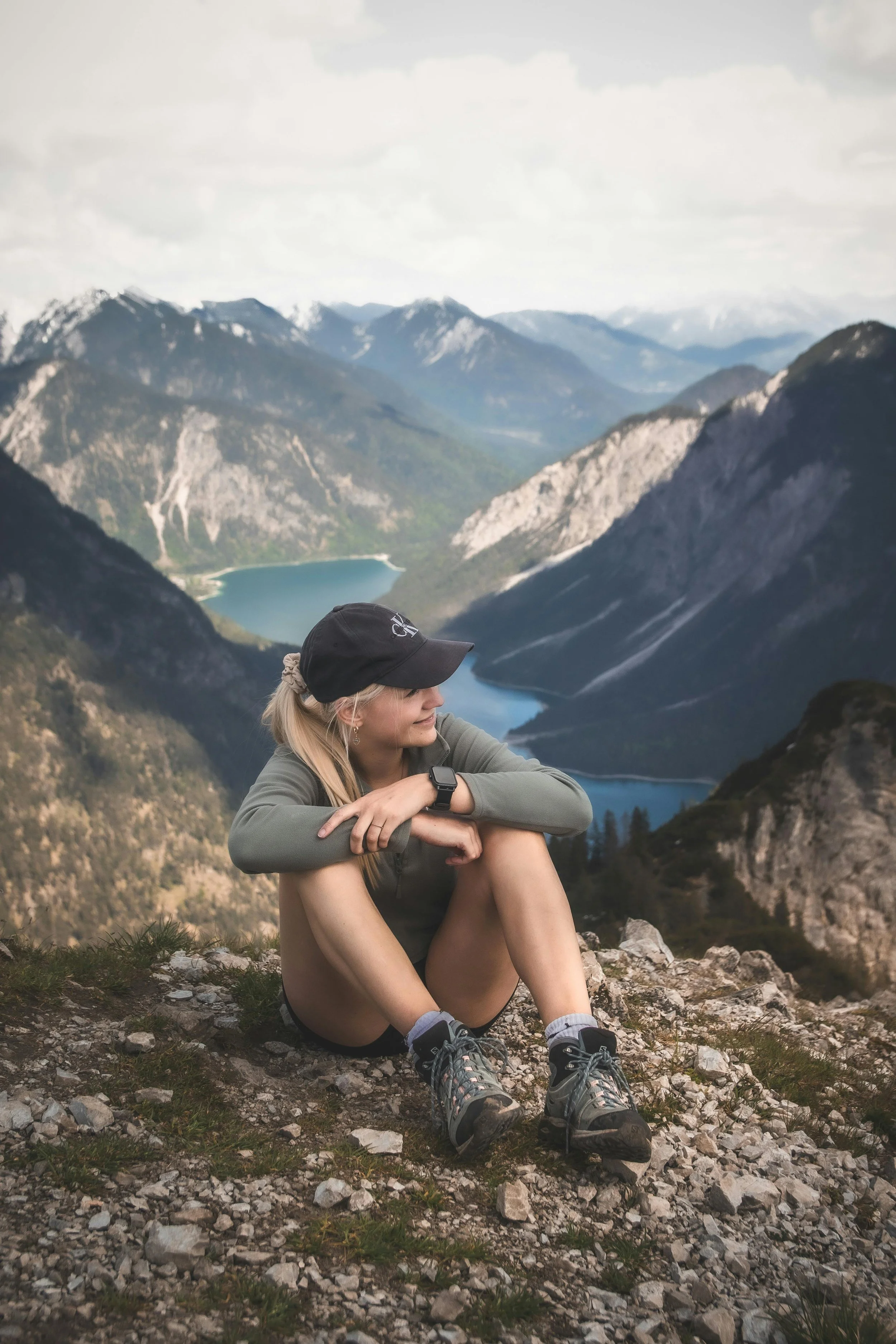 Woman in hiking gear sitting on a rocky mountain summit overlooking a glacial lake reflecting on her fitness journey and body wellness