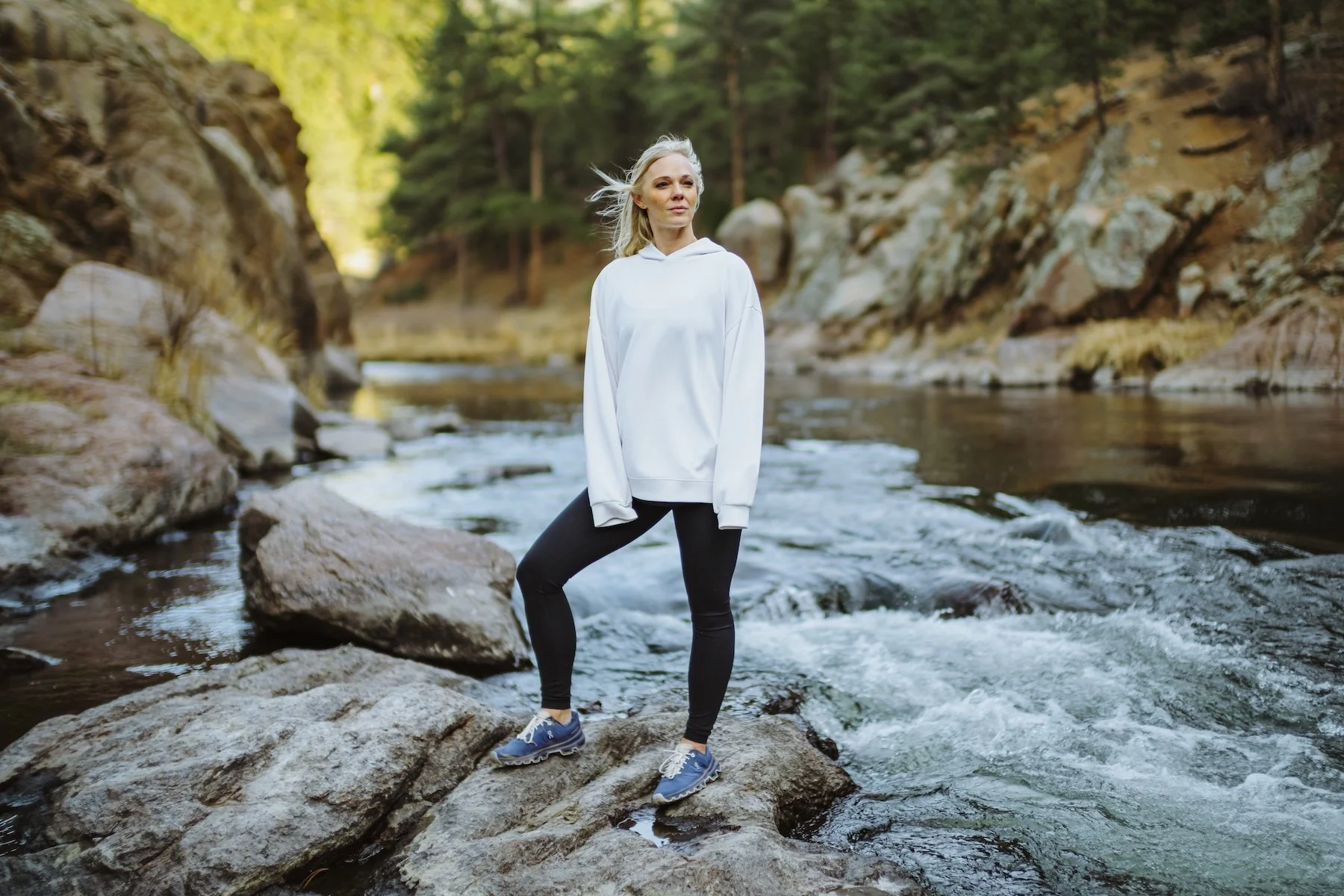 A woman standing on rocks in a river surrounded by a forested landscape.