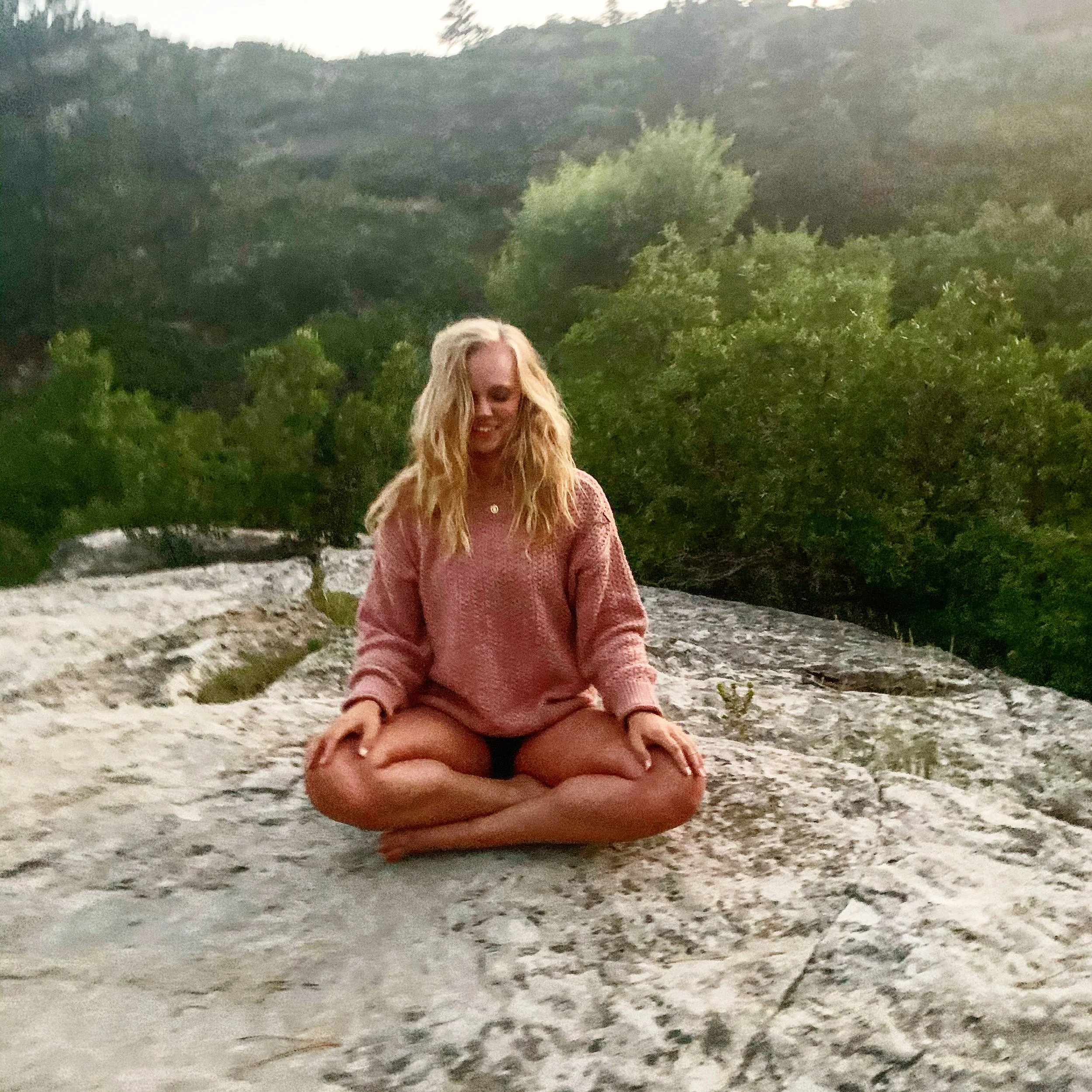 A woman with long blonde hair sitting cross-legged on a rock in a natural outdoor setting, wearing a pink sweater and black shorts, smiling with her eyes closed. Dense green trees and hills are in the background.