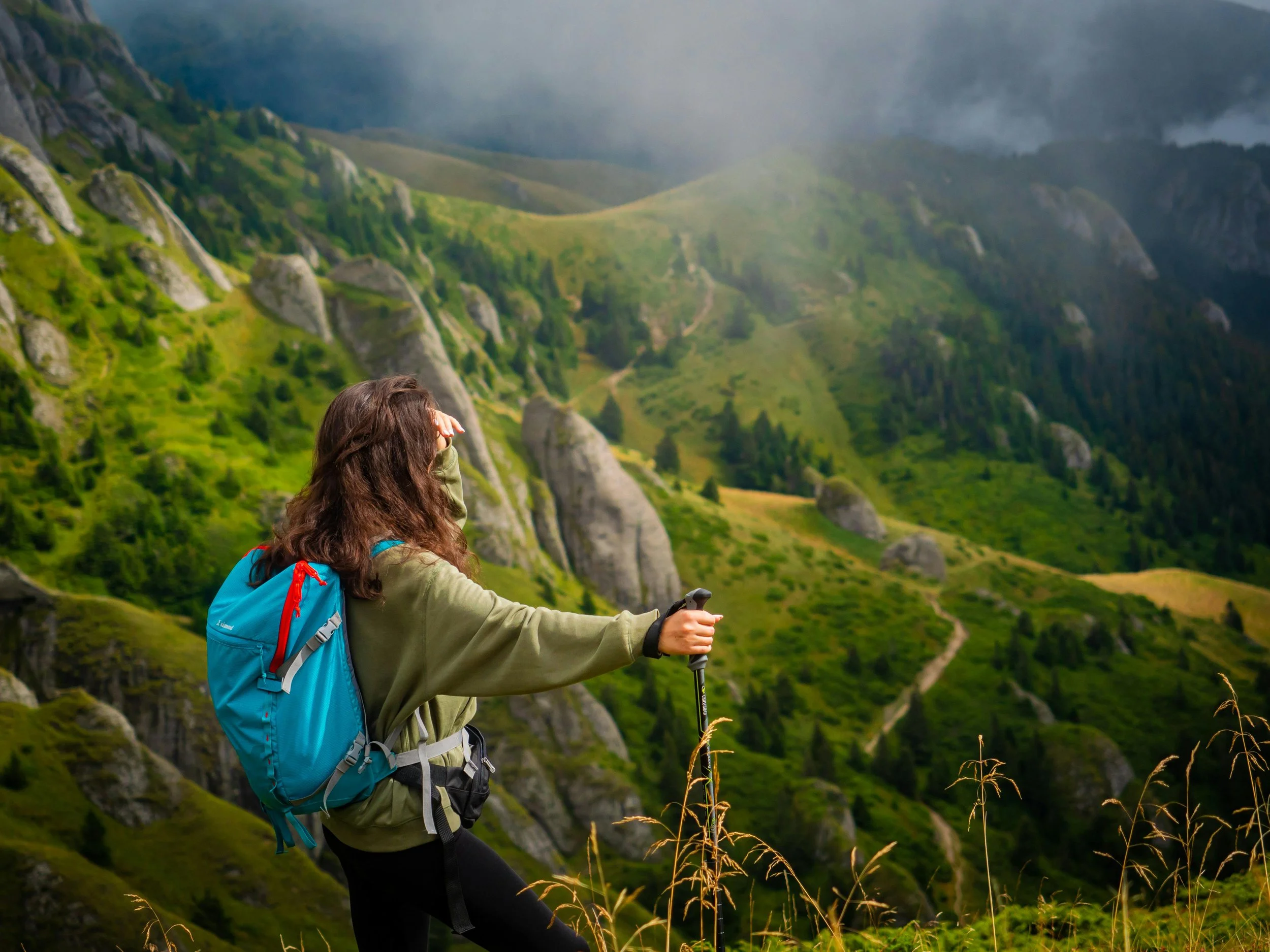 Woman with a hiking pack and trekking pole pausing on a mountain trail overlooking a green valley representing outdoor fitness and functional strength. Photo by Corneliu Stefan Esanu on Pexels