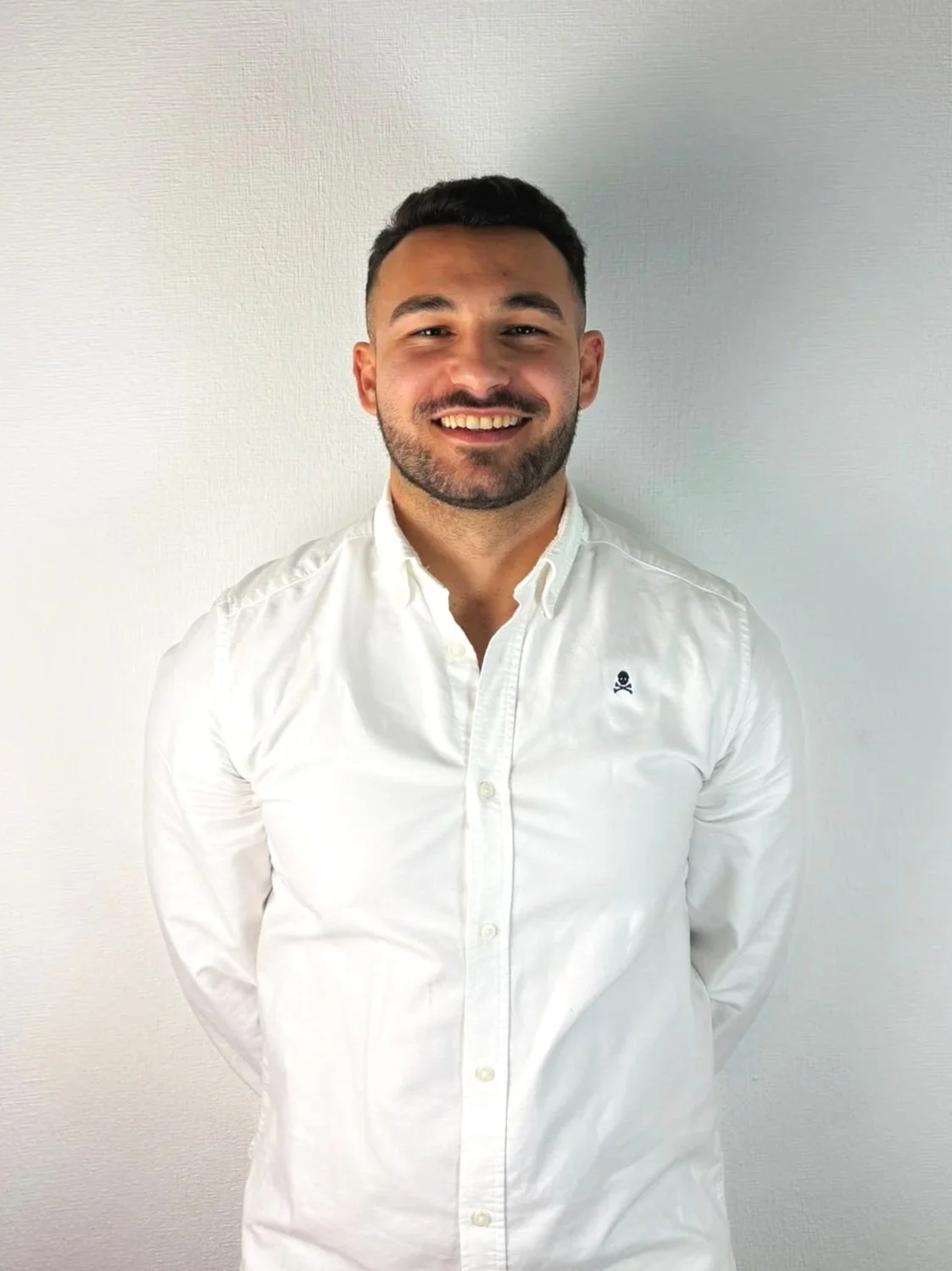 A young man with short dark hair and a beard, smiling, wearing a white button-up shirt with a small skull emblem on the left chest, standing against a plain light-colored wall.