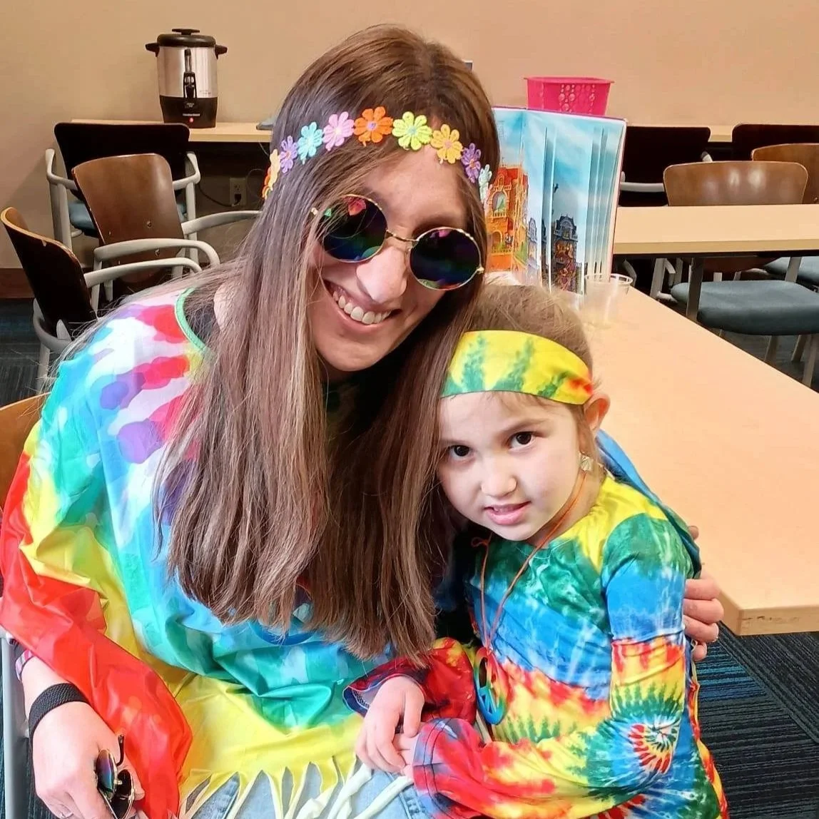 A woman and young girl dressed in colorful tie-dye shirts, with the woman wearing flower crown accessories and round sunglasses, and the girl wearing a tie-dye headband. They are smiling and hugging in a room with tables and chairs, and a coffee pot 