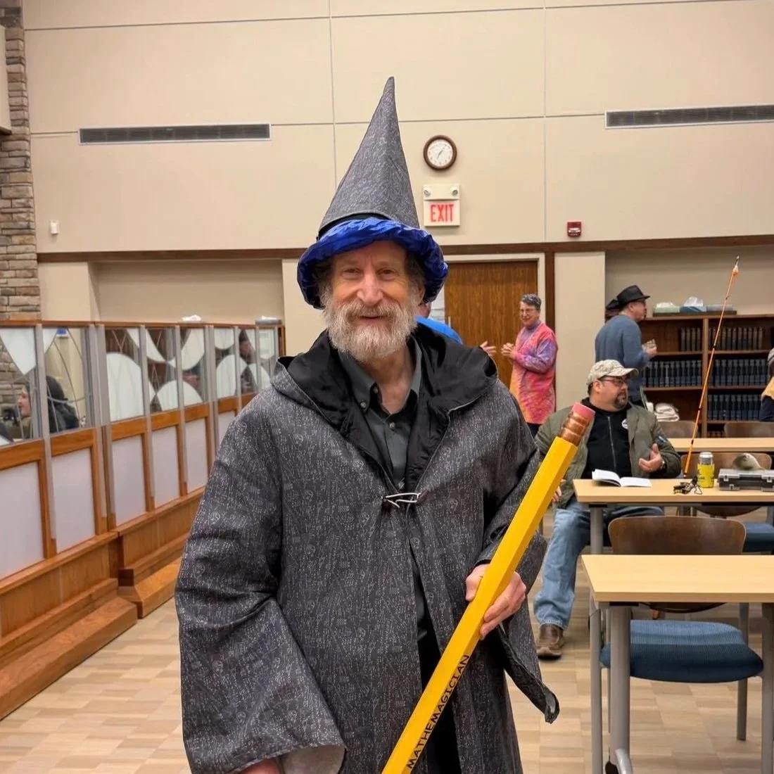 A man dressed as a wizard, wearing a tall pointed wizard hat and a cape, smiling and holding a large yellow pencil. The setting appears to be an indoor event or gathering with other people in the background.