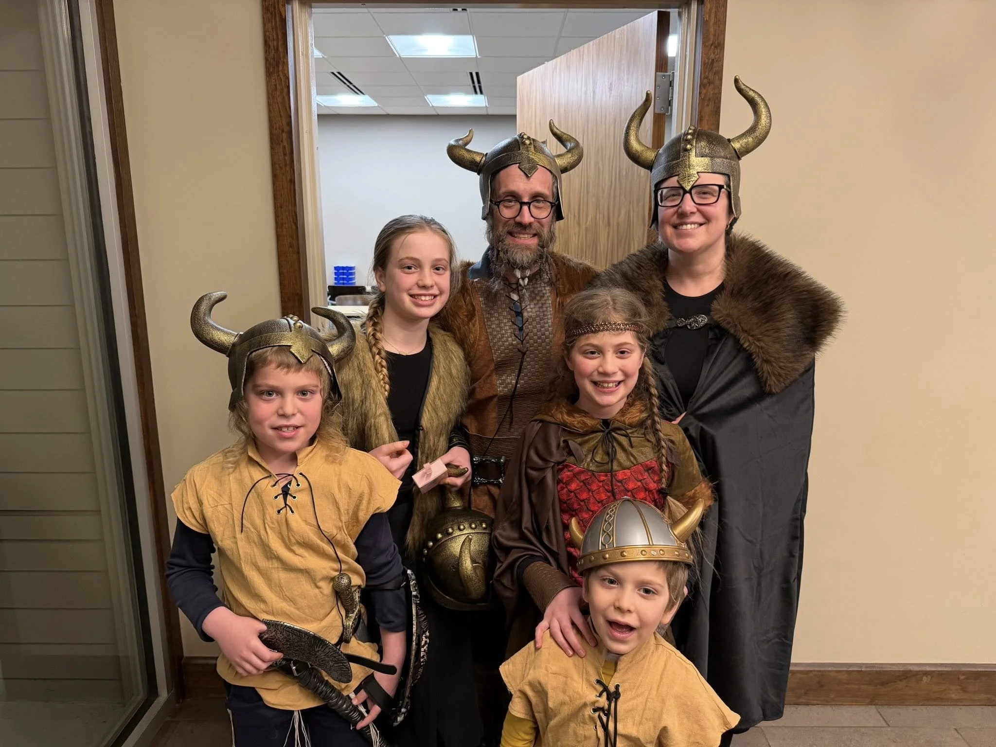 People dressed in Viking costumes, wearing horned helmets, standing in a room with beige walls and a wooden door.