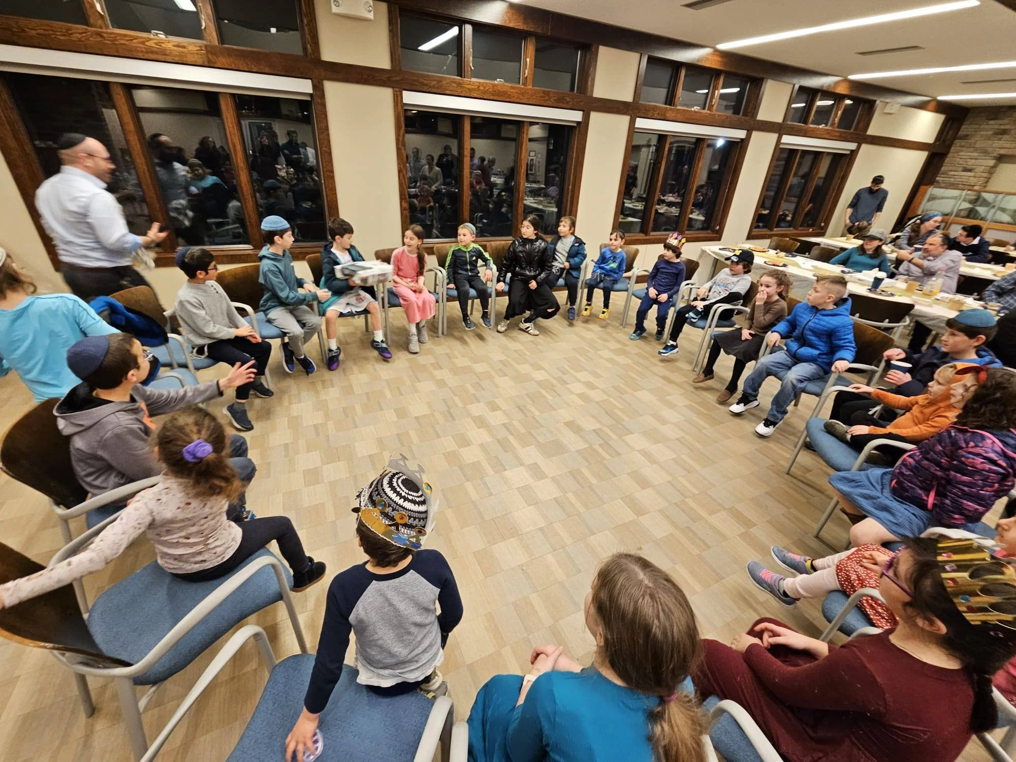A group of children sitting in a circle in a large room with big windows, participating in a group activity or discussion, while a man stands nearby talking.