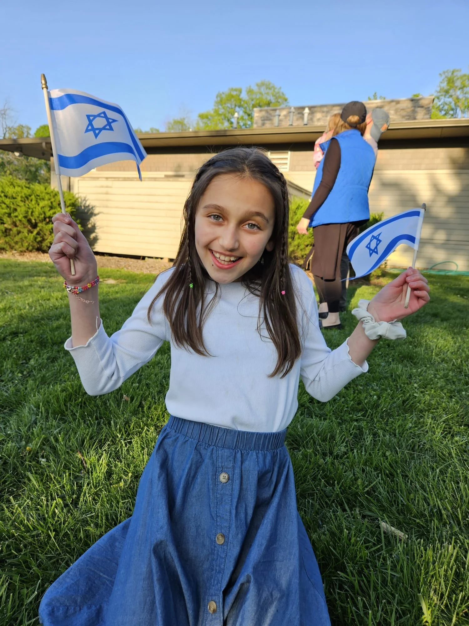 A young girl with long brown hair holding Israeli flags, smiling at the camera, standing on grass with a house and trees in the background.
