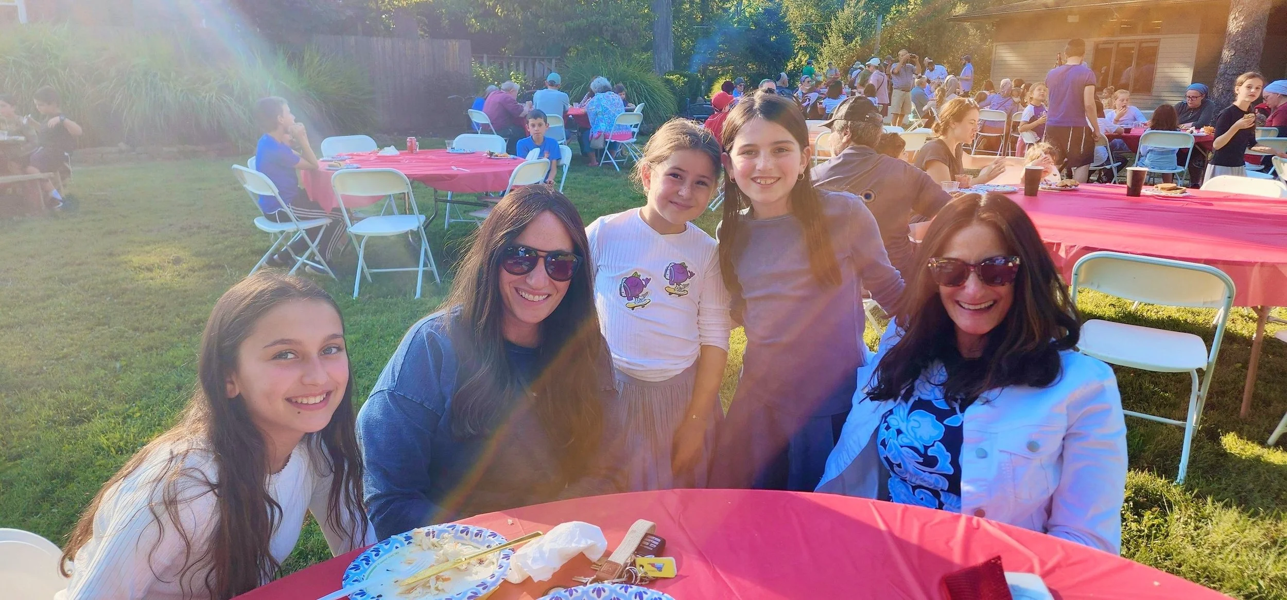 A group of five women and girls smiling and posing for a photo at an outdoor gathering or party in the sunlight. There are tables with red tablecloths and chairs, with many other people in the background sitting, standing, and socializing in a backyard or garden setting.