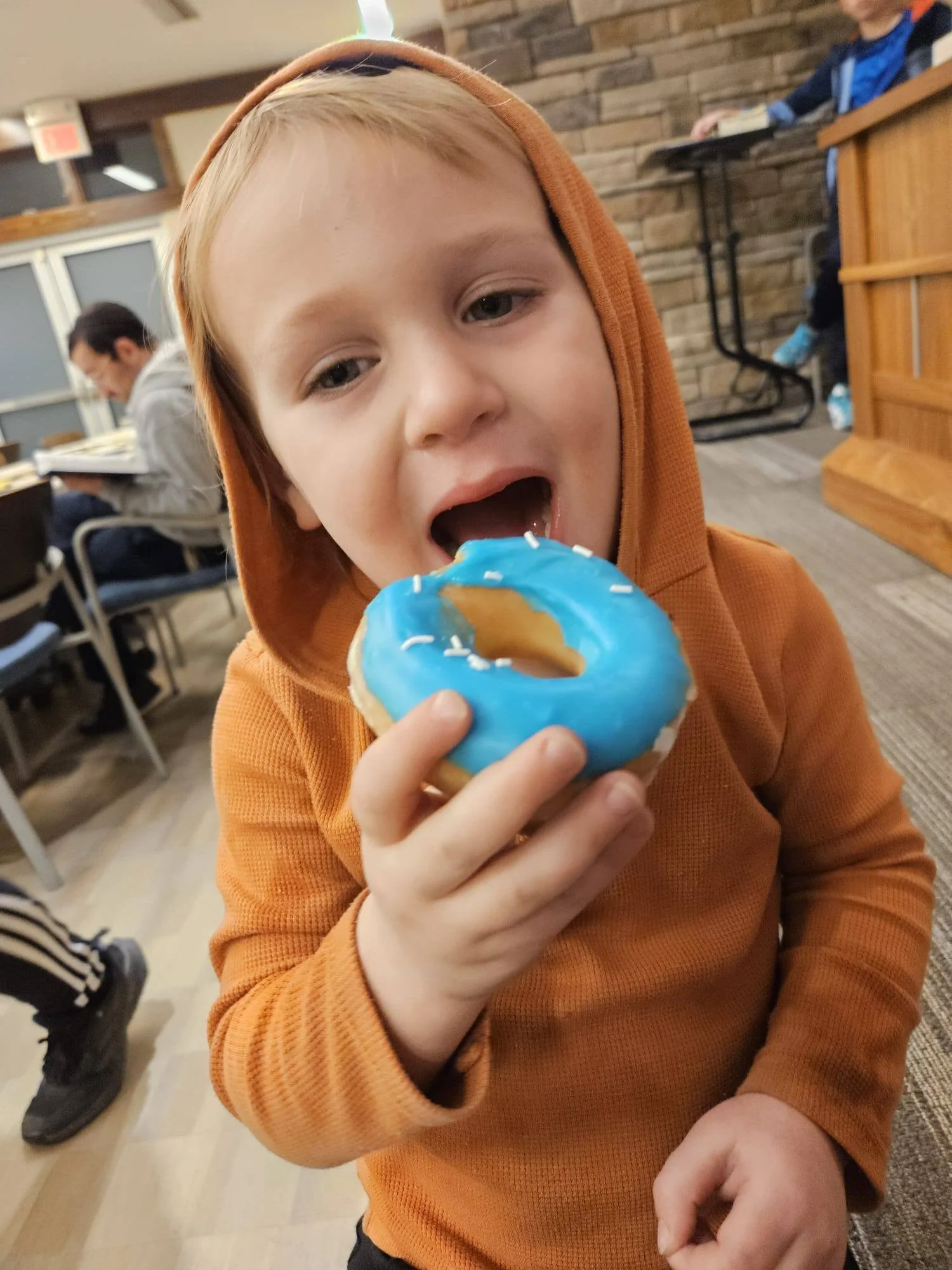 A young boy with light skin and blond hair, wearing an orange hoodie, is holding a donut with blue icing and white sprinkles, and is about to take a bite.
