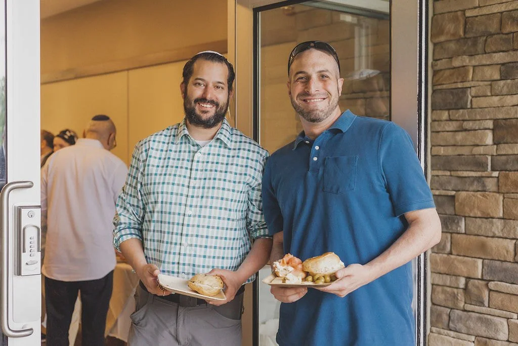 Two men smiling at the camera while holding plates of food at a social gathering, standing in front of a glass door with a stone wall on the side.