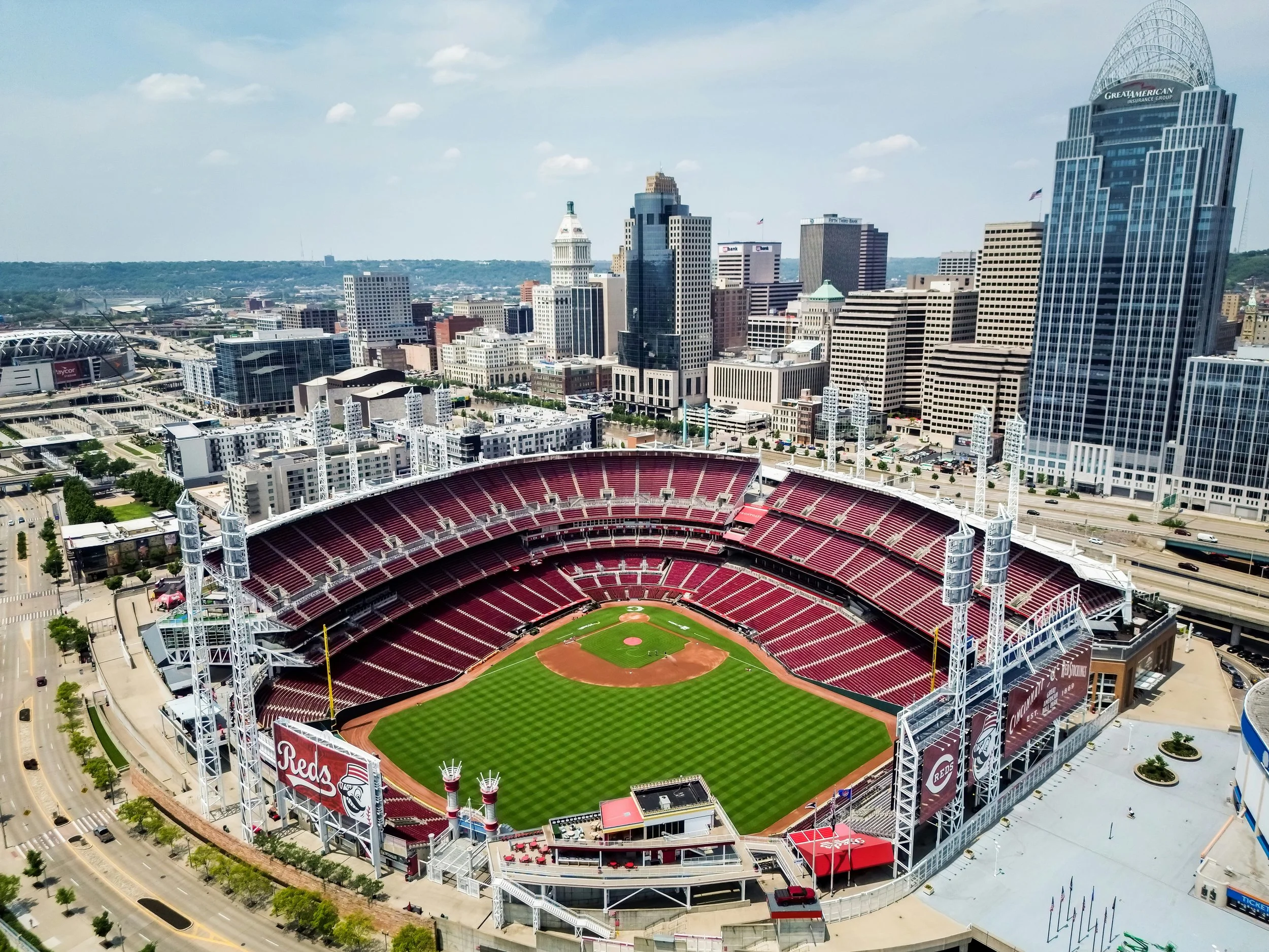 Aerial view of a baseball stadium with red seats, green field, and surrounding city skyline with tall skyscrapers under blue sky.