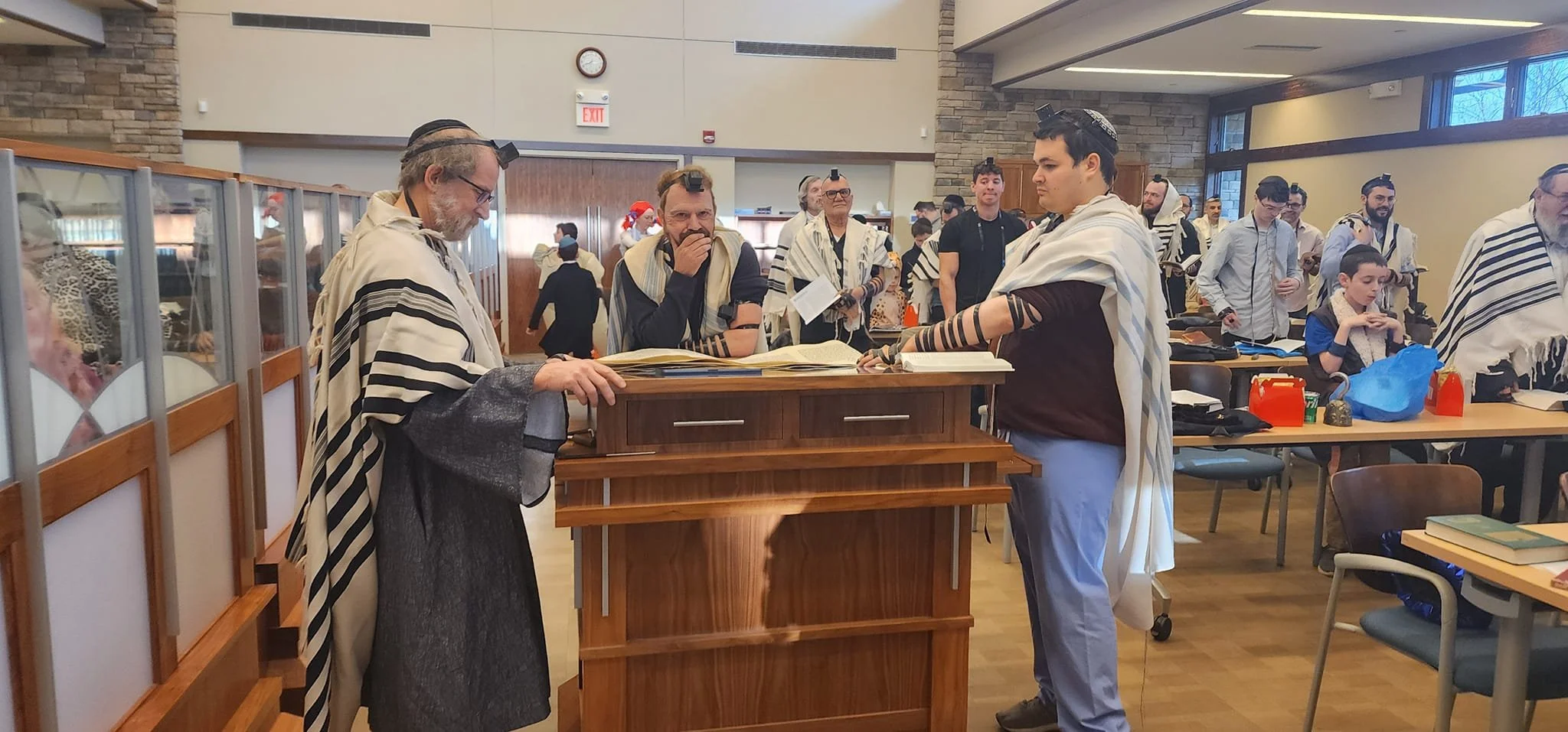 Group of people participating in a Jewish prayer service, standing around a wooden prayer podium in a synagogue. Men are wearing tallit prayer shawls and kippahs. Some individuals are engaged in prayer, reading from prayer books, or observing the ceremony.