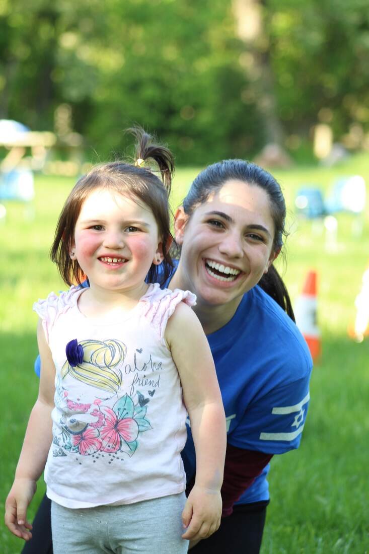 A young girl and a woman smiling outdoors in a park or backyard with green trees in the background.