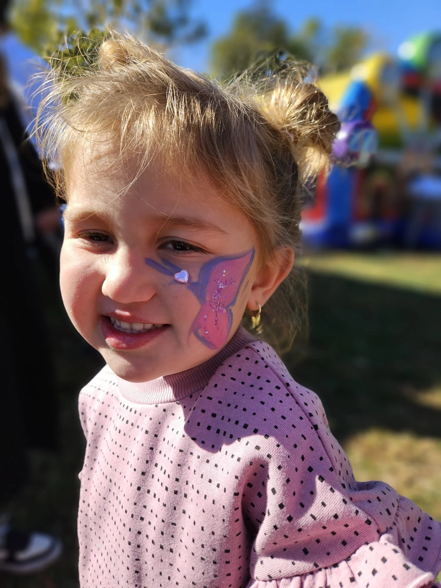 Close-up of a young girl with face paint of a butterfly, smiling outdoors on a sunny day, wearing a pink polka dot shirt.