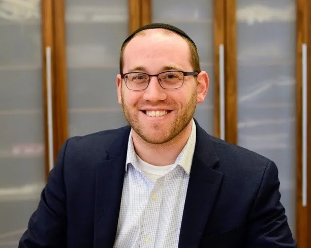 A smiling man with glasses, wearing a suit jacket and a collared shirt, sitting in front of wooden cabinets.