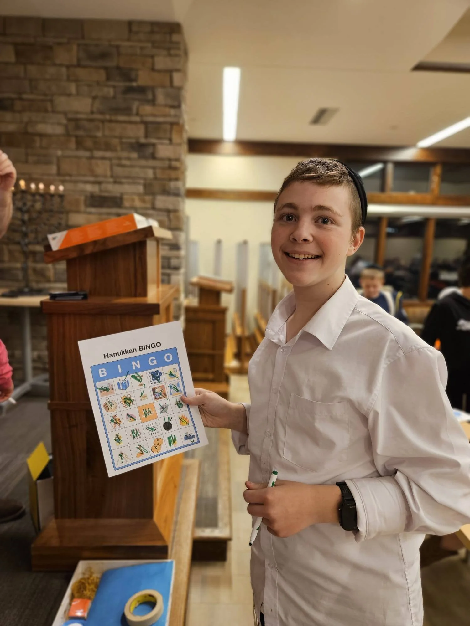 Young man with short hair smiling, holding a green marker in his right hand, standing in a room with wooden shelves and a brick wall, holding a Hanukkah Bingo game card.