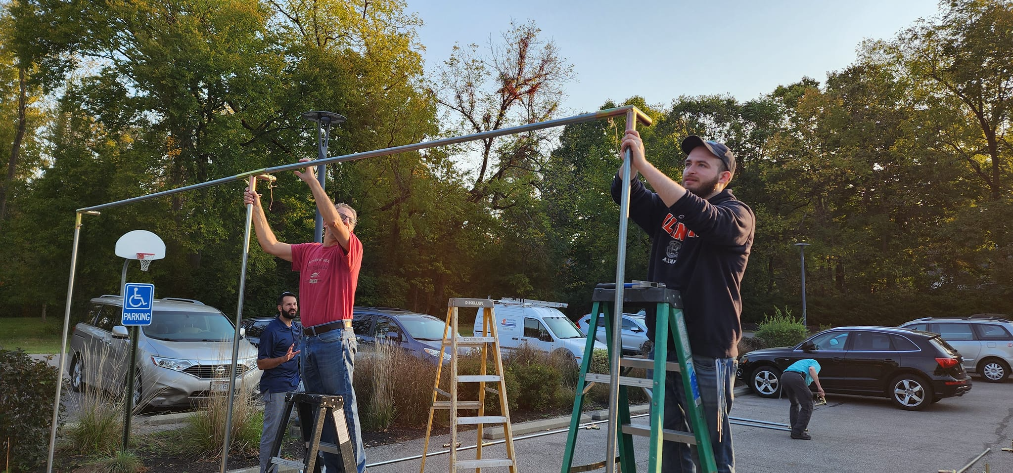 Two men setting up a basketball hoop outdoors with a parking lot and trees in the background.
