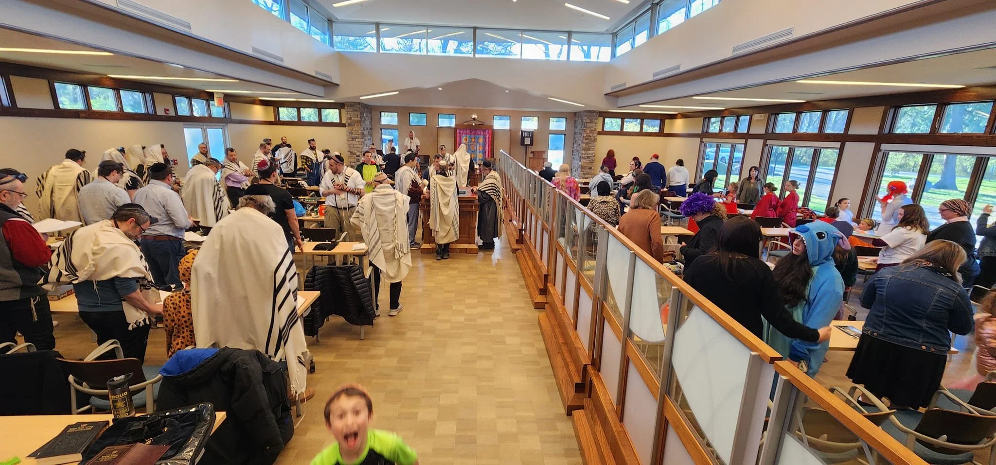 A large indoor gathering with many people, some dressed in traditional Jewish garments, in a spacious, well-lit room with large windows and wooden accents.