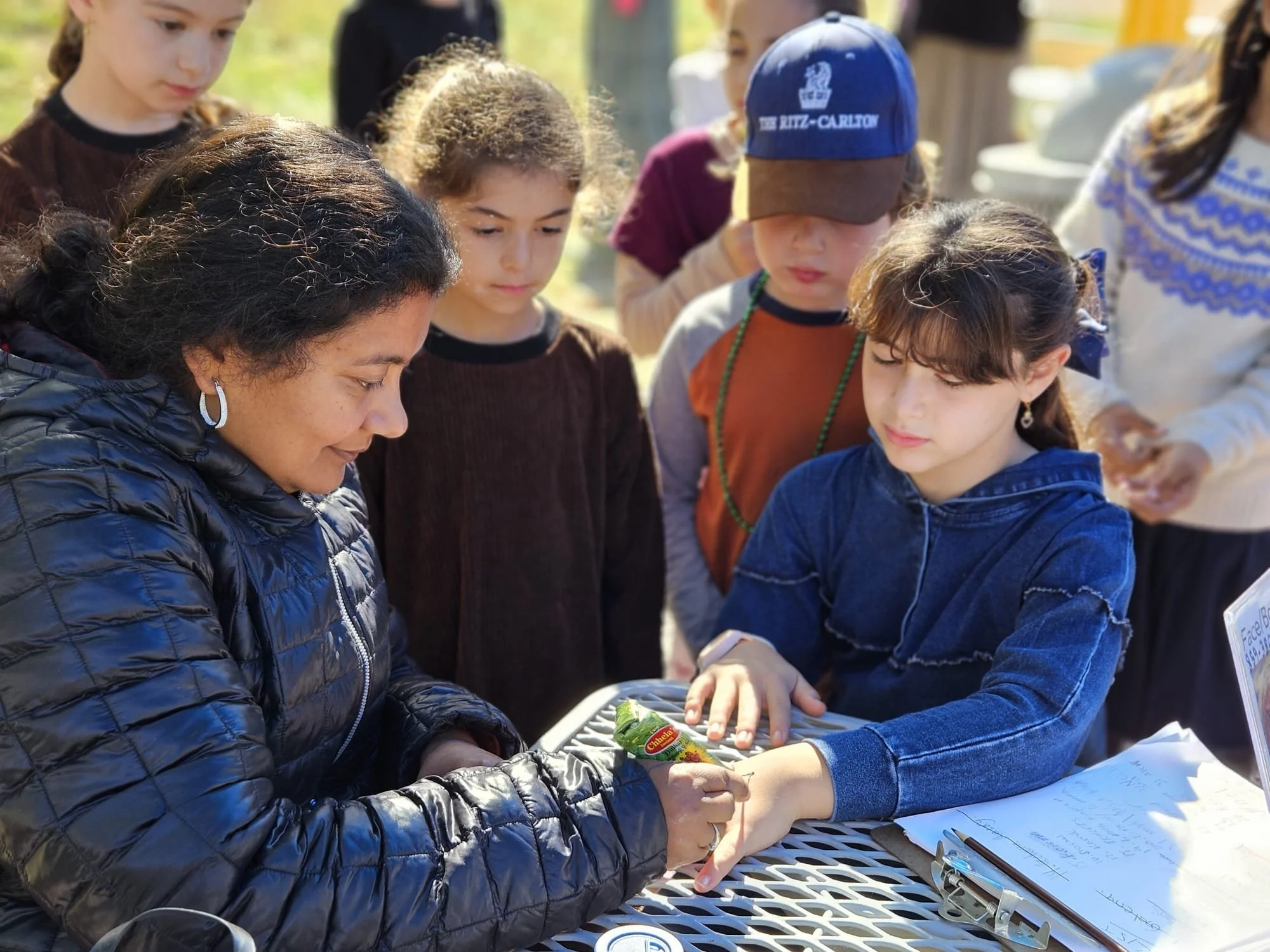 A group of children and an adult gathered around a table outdoors, examining something together. The children are looking at a tray with a plant or vegetables, with notes and a pen nearby, suggesting an educational activity or workshop.