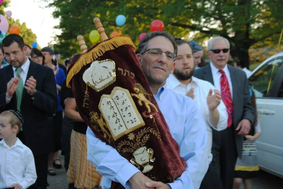 A man holding a Torah scroll during a religious celebration, surrounded by a group of people clapping, outdoors with trees and colorful balloons in the background.