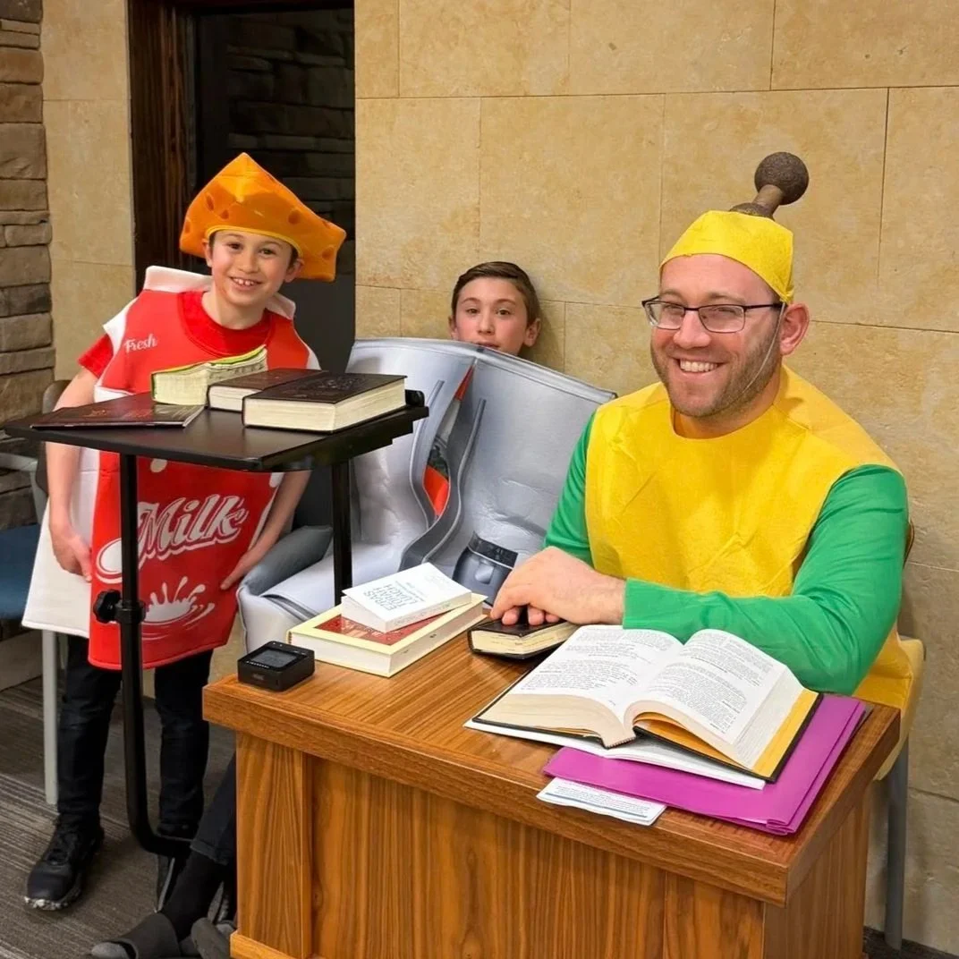 Three people dressed in cheese-themed costumes, with two children and an adult man, sitting at a table with books and papers in a room with beige walls.