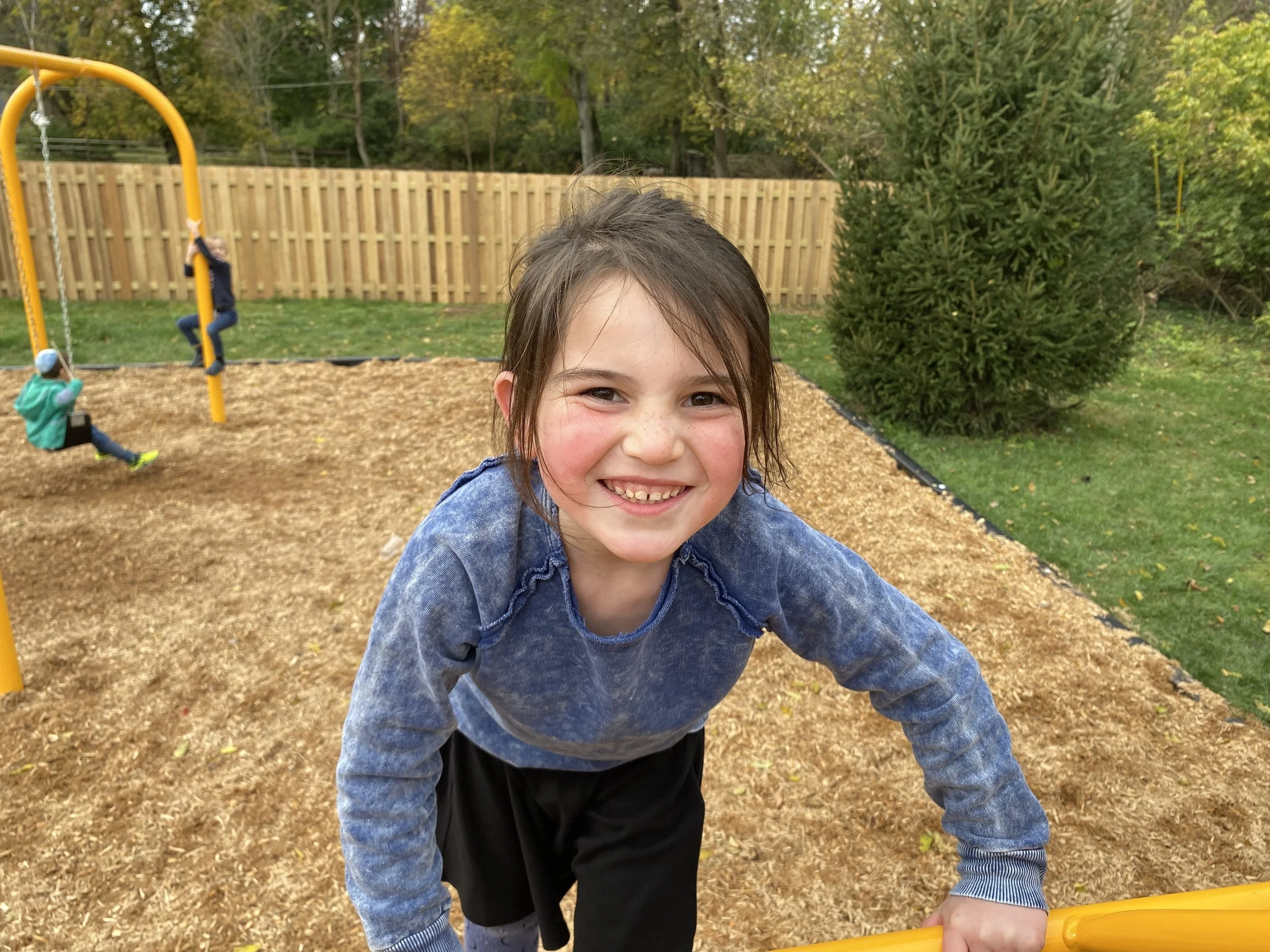 Young girl smiling at the camera in a park with swings and trees in the background.