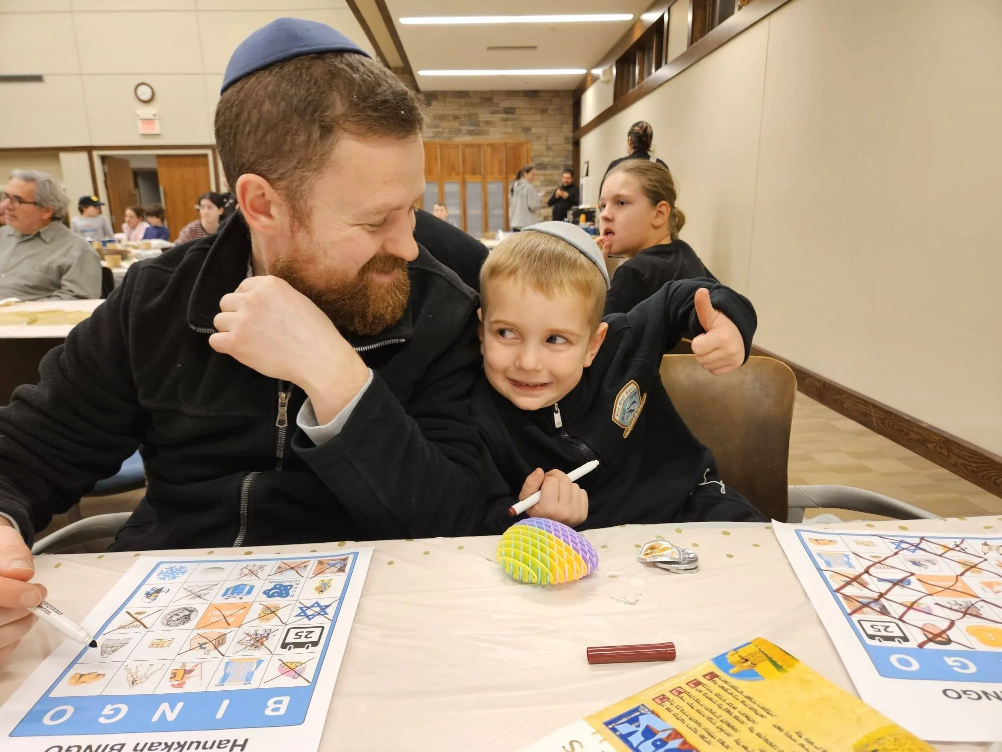 A man with a beard and a young boy wearing a kippah are sitting at a table playing bingo. The man is smiling and leaning toward the boy, who is holding a marker. Other people are seated at tables and appear to be engaged in an activity, with some sta