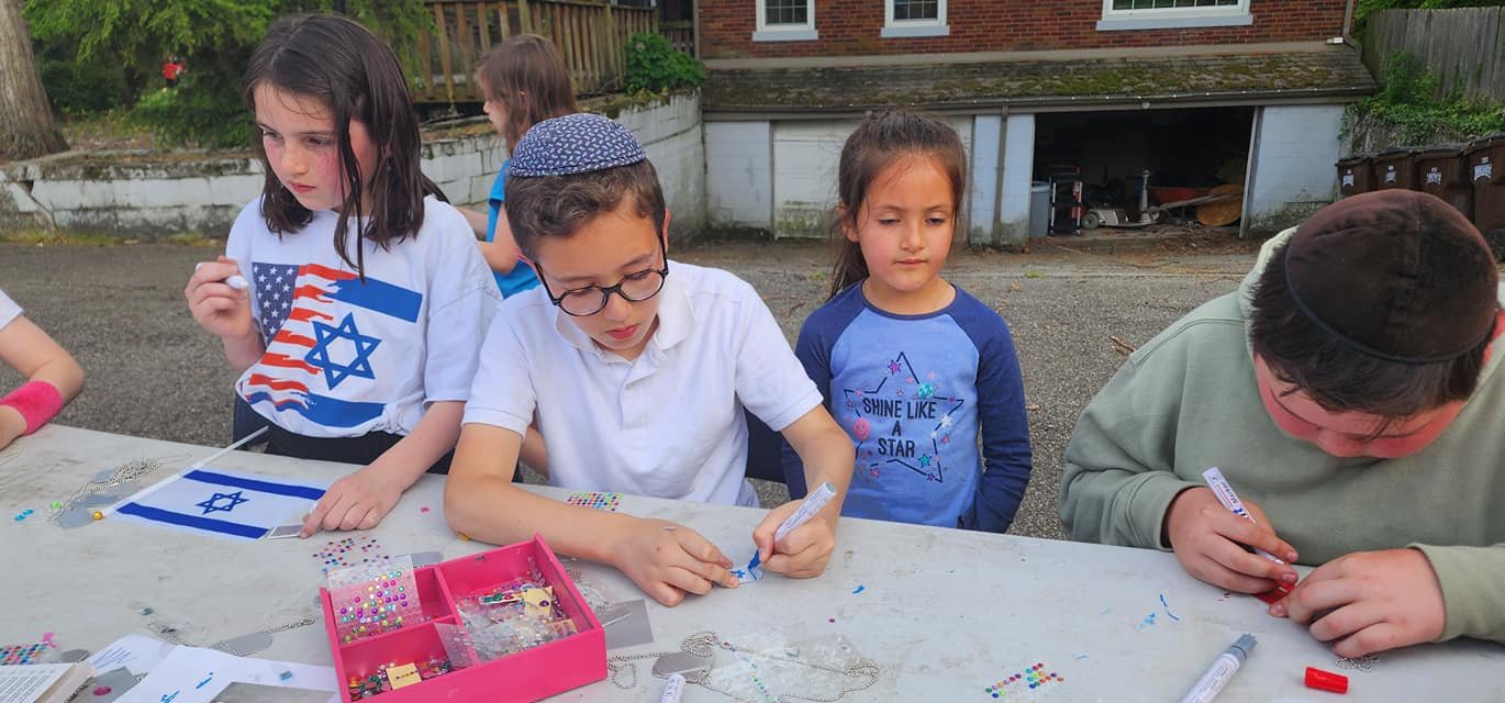 Children decorate small Israeli flags with beads and markers at a table outdoors.