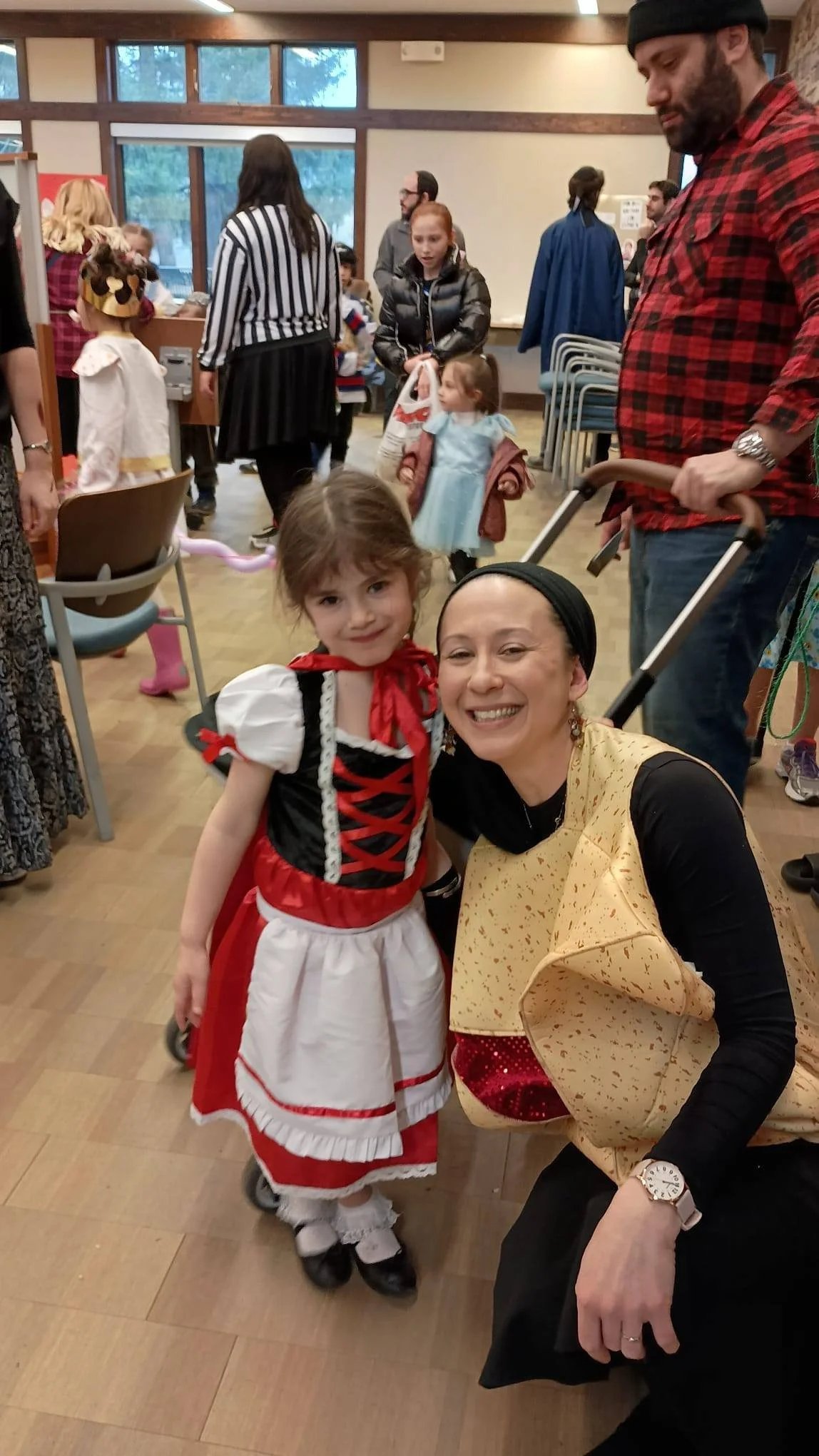 A woman and a young girl in costumes, smiling at the camera at a costume party or event. The woman is kneeling, wearing a black top with a yellow cape and a red and yellow cape, while the girl is dressed in a red and white dress with black shoes and white socks. People in costumes and casual clothing are in the background, inside a room with large windows.