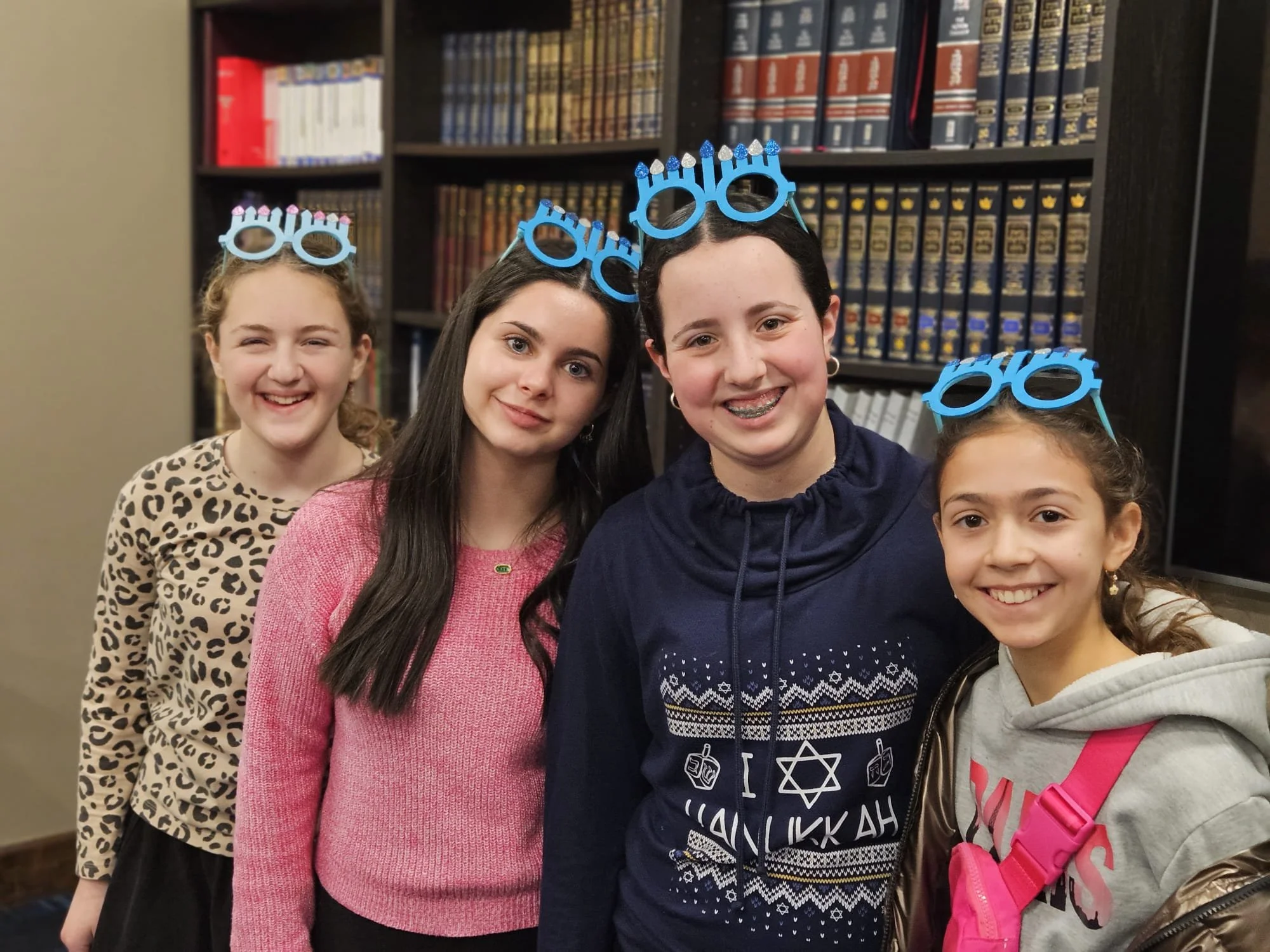 Four children, three girls and one boy, wearing blue candle holders with candles as holiday headbands, smiling and standing in front of a bookshelf filled with books.