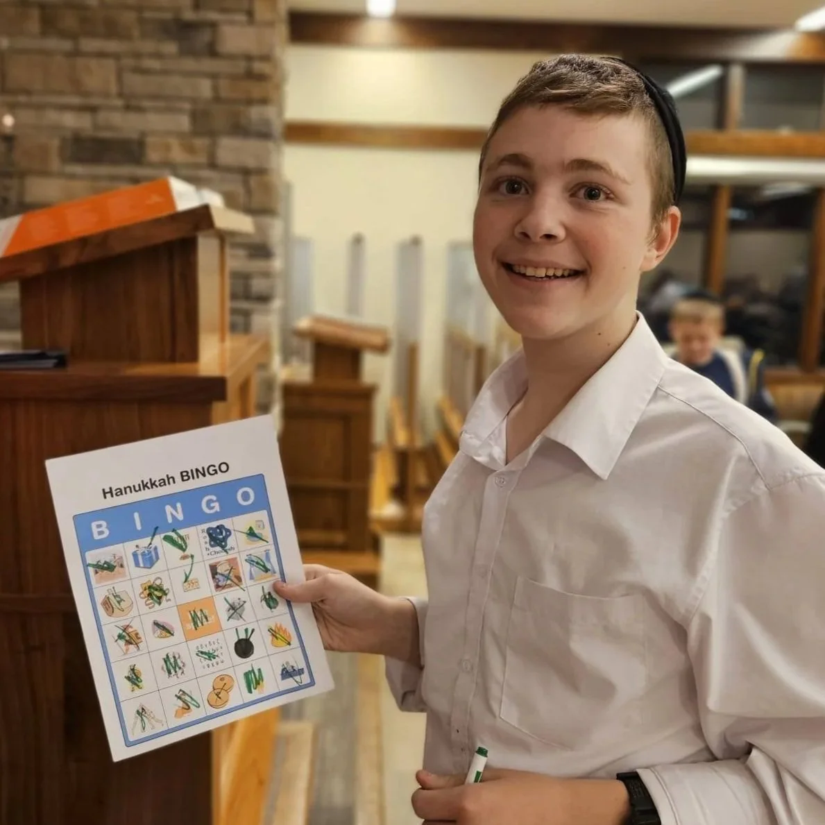 A young boy smiling at the camera, holding a Hanukkah Bingo card with Hebrew symbols and holiday-themed images, standing in a room with wooden furniture.