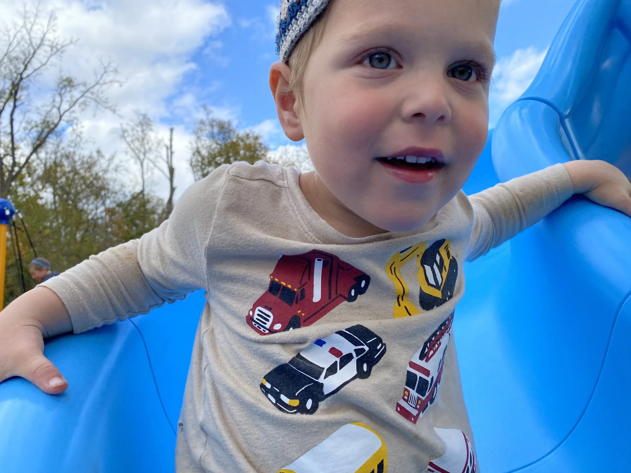 A young boy with blue eyes and light skin leaning on a blue plastic surface outdoors, wearing a gray and blue knit hat and a T-shirt with colorful vehicle graphics, in a park with trees and a partly cloudy sky in the background.