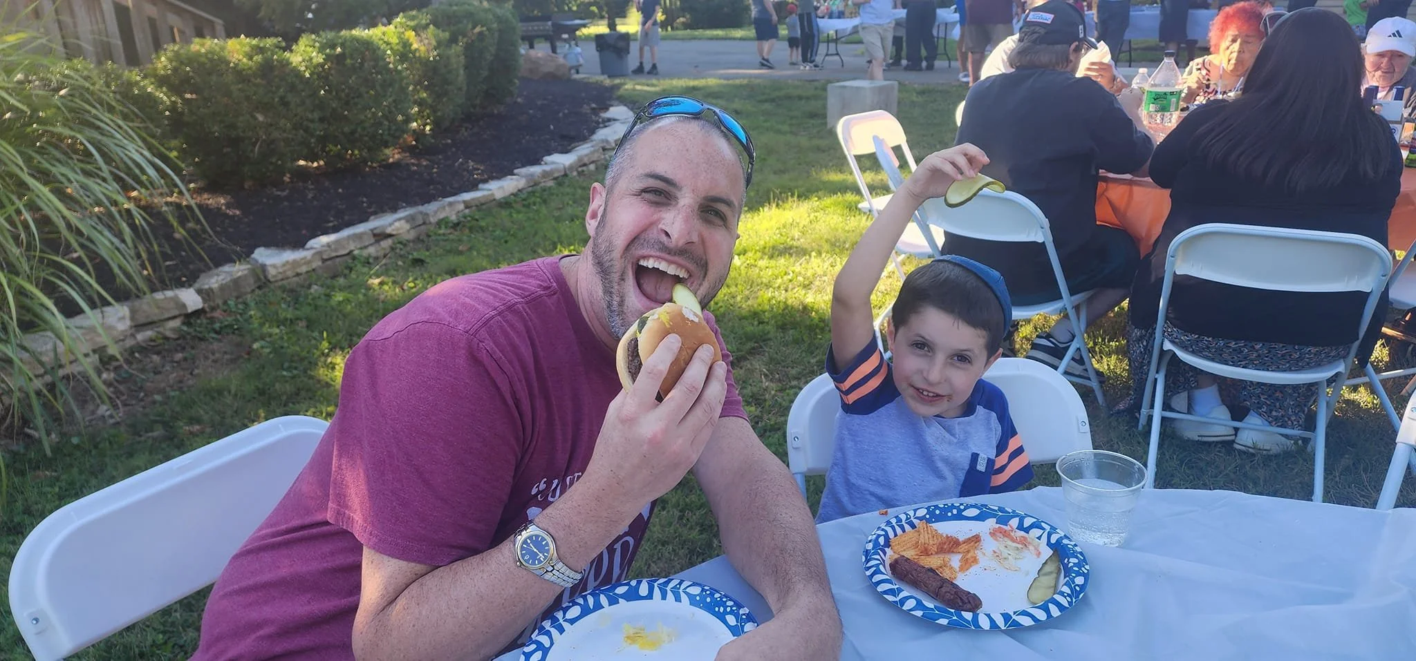 A man and a young boy sitting at a table outdoors during a gathering. The man is holding a hot dog and smiling at the camera, while the boy is also holding a hot dog and smiling. There are paper plates with food and a clear plastic cup on the table. Other people are sitting at tables in the background.