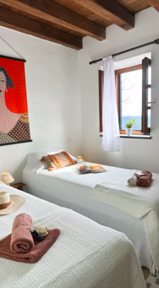 Two clean, white beds with rolled towels and pillows in a bright bedroom with wooden beams ceiling, a window with white curtains, a small potted plant, and a colorful portrait hanging on the wall.