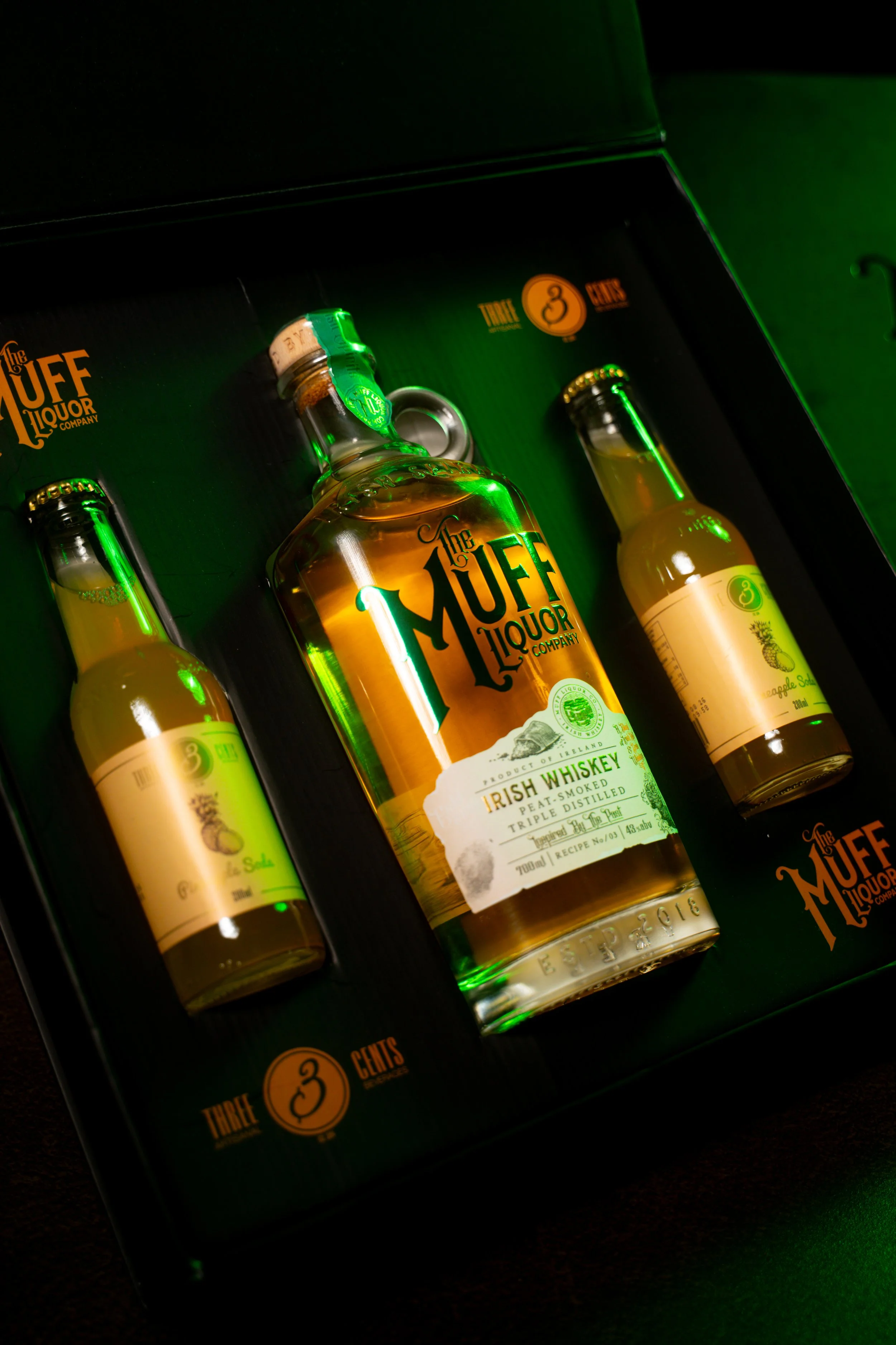 Set of three small bottles of flavored Irish whiskey from the Muff Liquor Company, packaged in a green box with black interior. The central bottle is clear with a white label, and the other two smaller bottles are also labeled and contain a yellowish liquid.