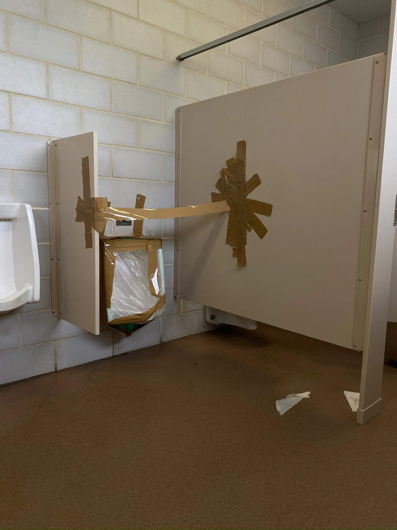 A bathroom stall with a door that is taped shut with brown tape to prevent use. A no toilet paper sign is visible on the wall beside the stall.